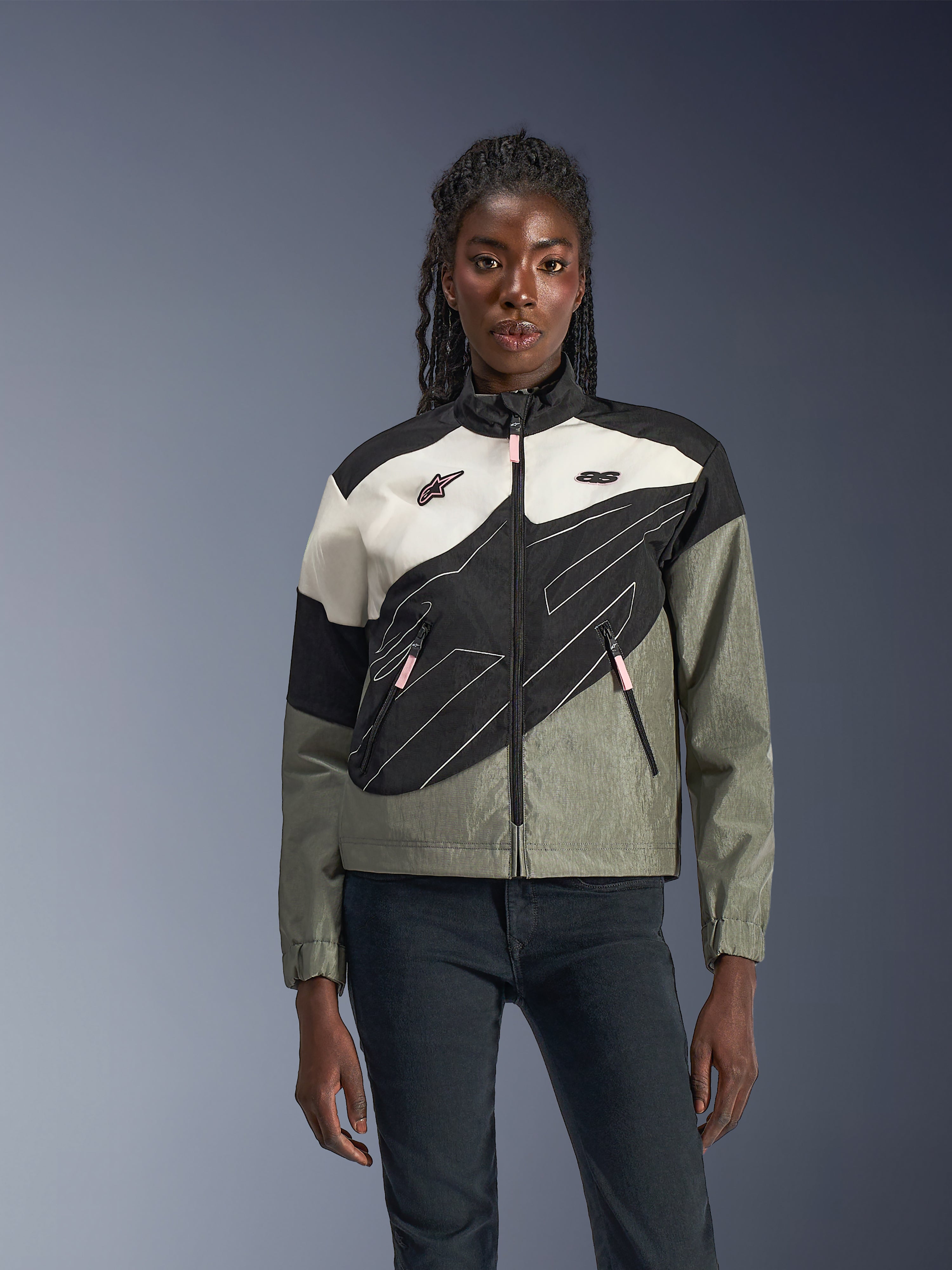 Vrouw die de Alpinestars Stella Moshell Jas draagt, urbane motorjas in Black Fur, grijs, gebroken wit en roze, asymmetrisch ontwerp met technische graphics, roze ritstrekkers en Alpinestars-logo's, eigentijdse streetwear-stijl voor stedelijke rijders.