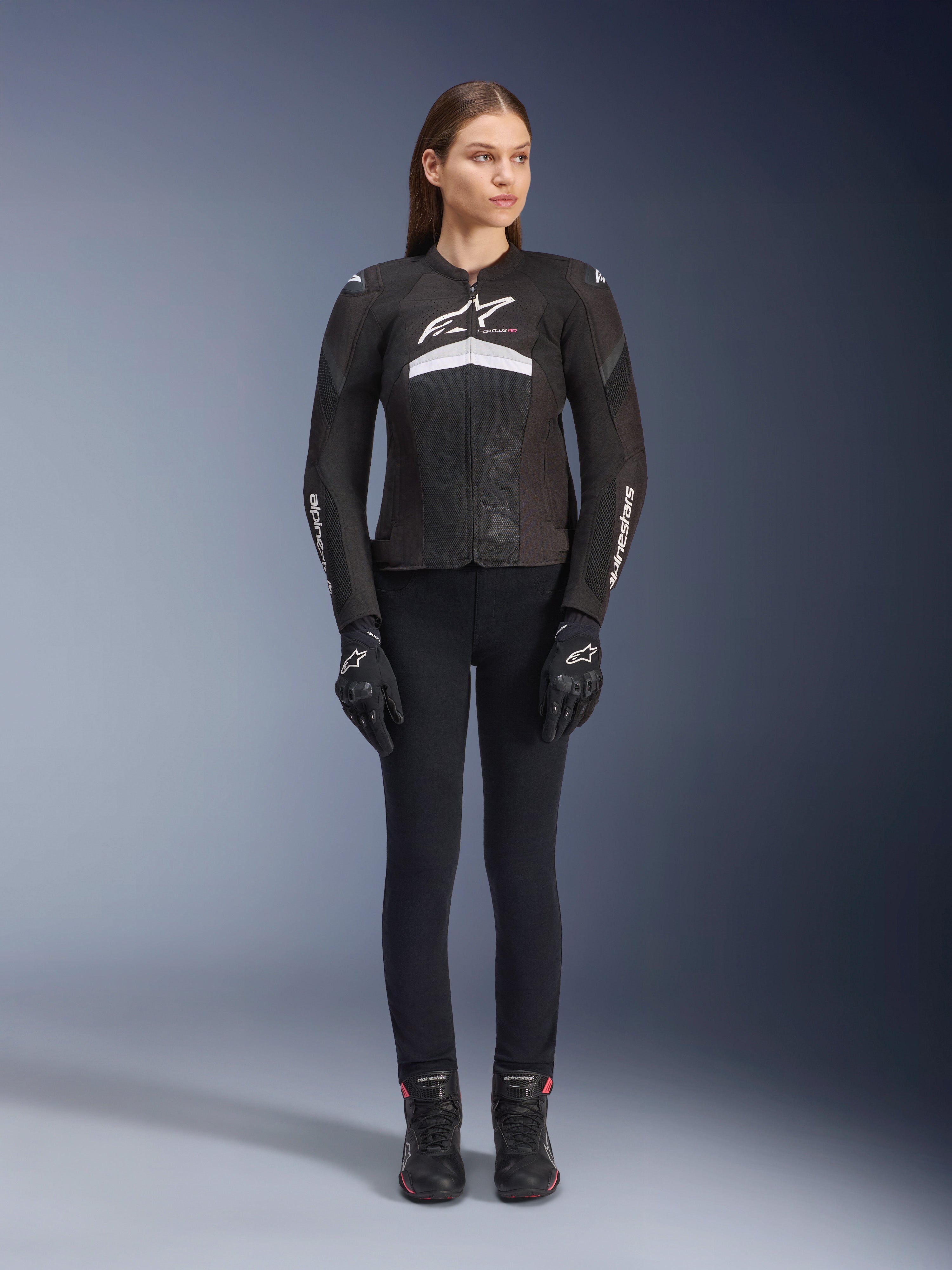 Vrouwelijke motorrijder draagt de Stella T-GP Plus R V4 Airflow Jas, zwart en wit, gecombineerd met een zwarte motorbroek, handschoenen en laarzen, voorzien van mesh-panelen voor ventilatie en witte Alpinestars-branding op de borst en armen