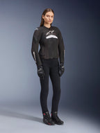 Vrouwelijke rijder in het Alpinestars Stella T-GP Plus R V4 Airflow Jacket, zwart en wit, met mesh-panelen voor ventilatie en witte logo's, gecombineerd met een zwarte broek, zwarte handschoenen en zwarte laarzen met roze accenten, sportieve motoruitrusting voor dames
