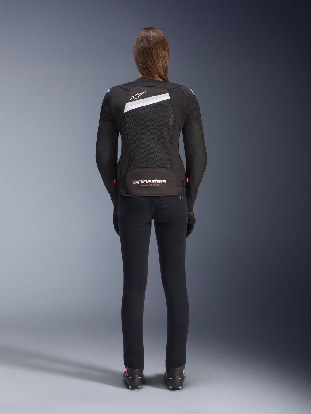Alpinestars Stella T-GP Plus R V4 Airflow Jacket, zwart en wit, achteraanzicht met uitgebreide mesh ventilatiepanelen en Alpinestars-branding, gedragen door een vrouwelijke rijder met een slim-fit zwarte motorbroek en laarzen, sportieve textiele motoruitrusting voor dames