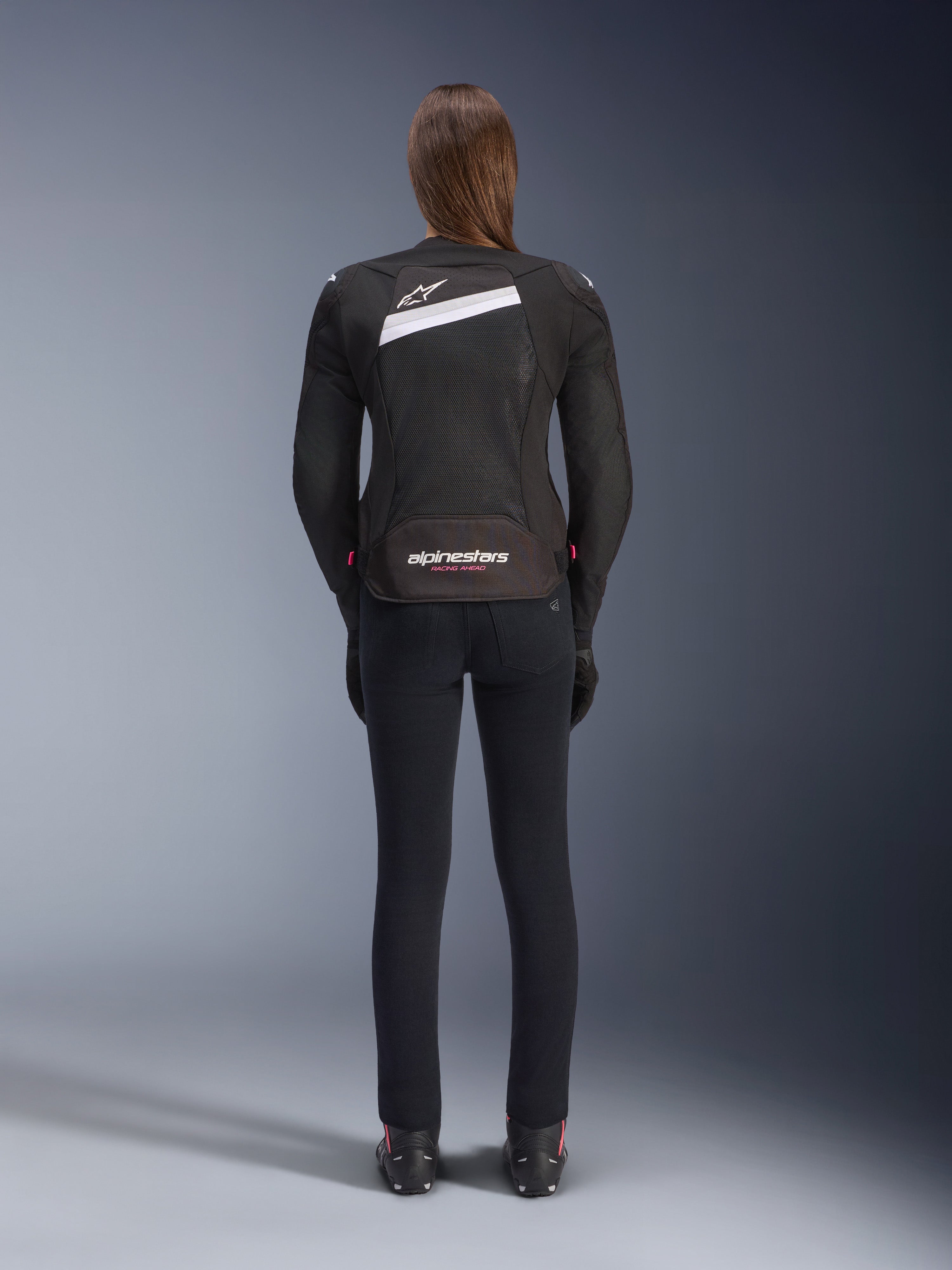 Alpinestars Stella T-GP Plus R V4 Airflow Jacket, zwart en wit, achteraanzicht met uitgebreide mesh ventilatiepanelen en Alpinestars-branding, gedragen door een vrouwelijke rijder met een slim-fit zwarte motorbroek en laarzen, sportieve textiele motoruitrusting voor dames