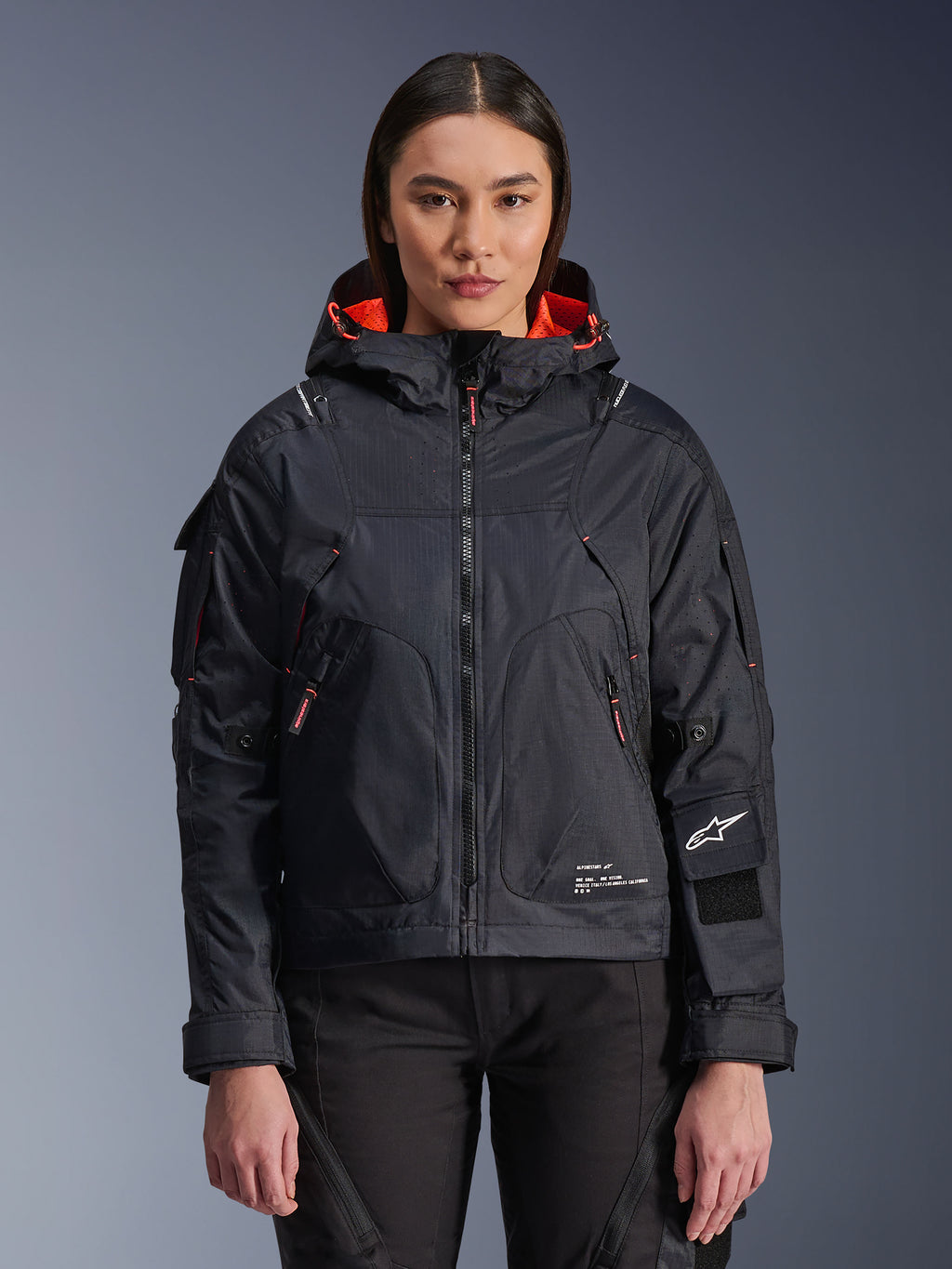 Alpinestars Molly Air Jacket, urban motorjas voor dames, zwart met Fluo Coral binnenvoering, gedragen door een vrouwelijk model, ontwerp met capuchon en technische textielconstructie, zakken met rits en Alpinestars-logo op de mouw