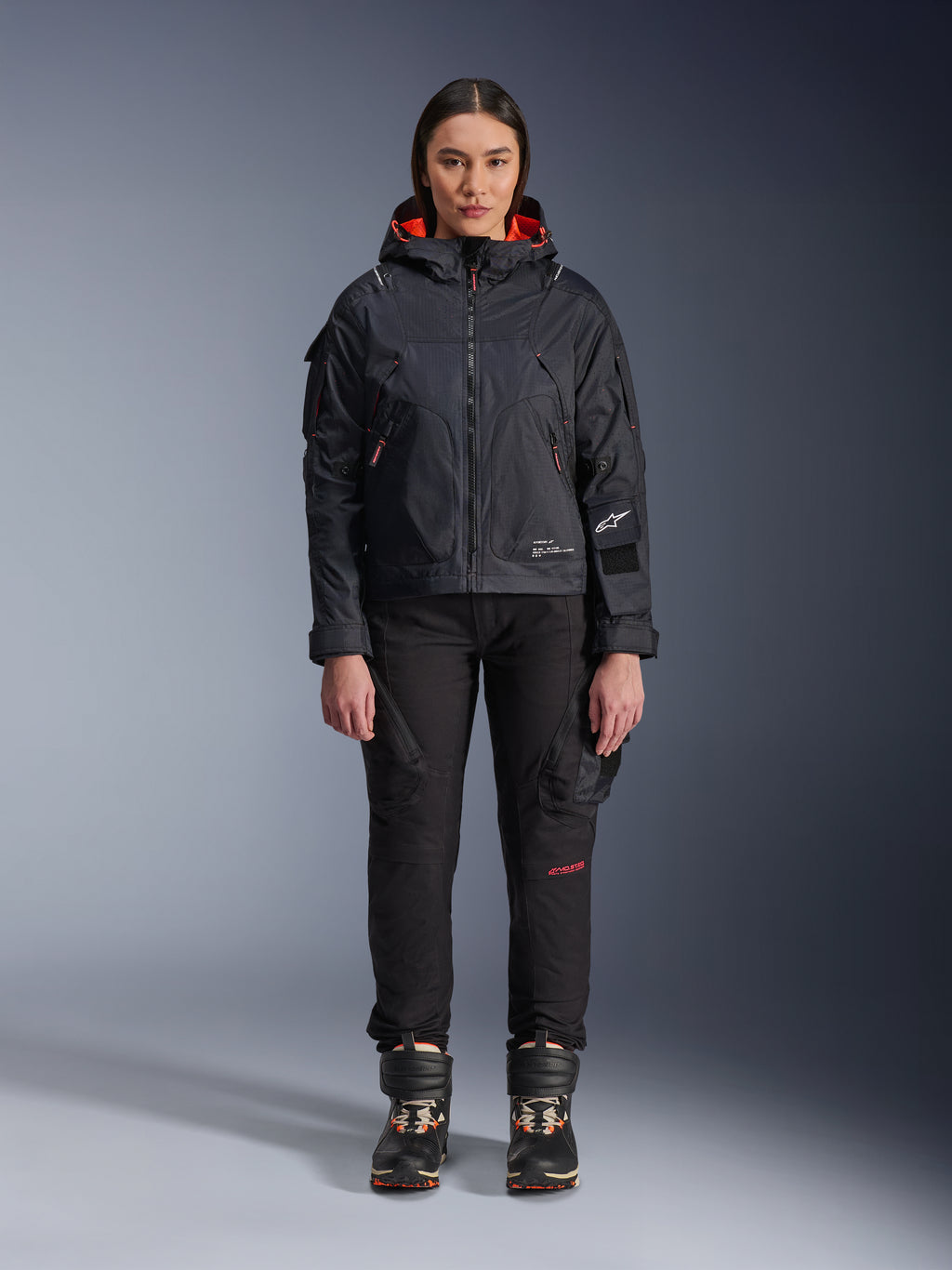 Vrouwelijk model draagt de Alpinestars Molly Air Jacket in Black en Fluo Coral, voorzien van een op utility geïnspireerd ontwerp met een capuchon en koraalrode binnenvoering, gecombineerd met een zwarte motorbroek en motorlaarzen, urban contemporary motoruitrusting