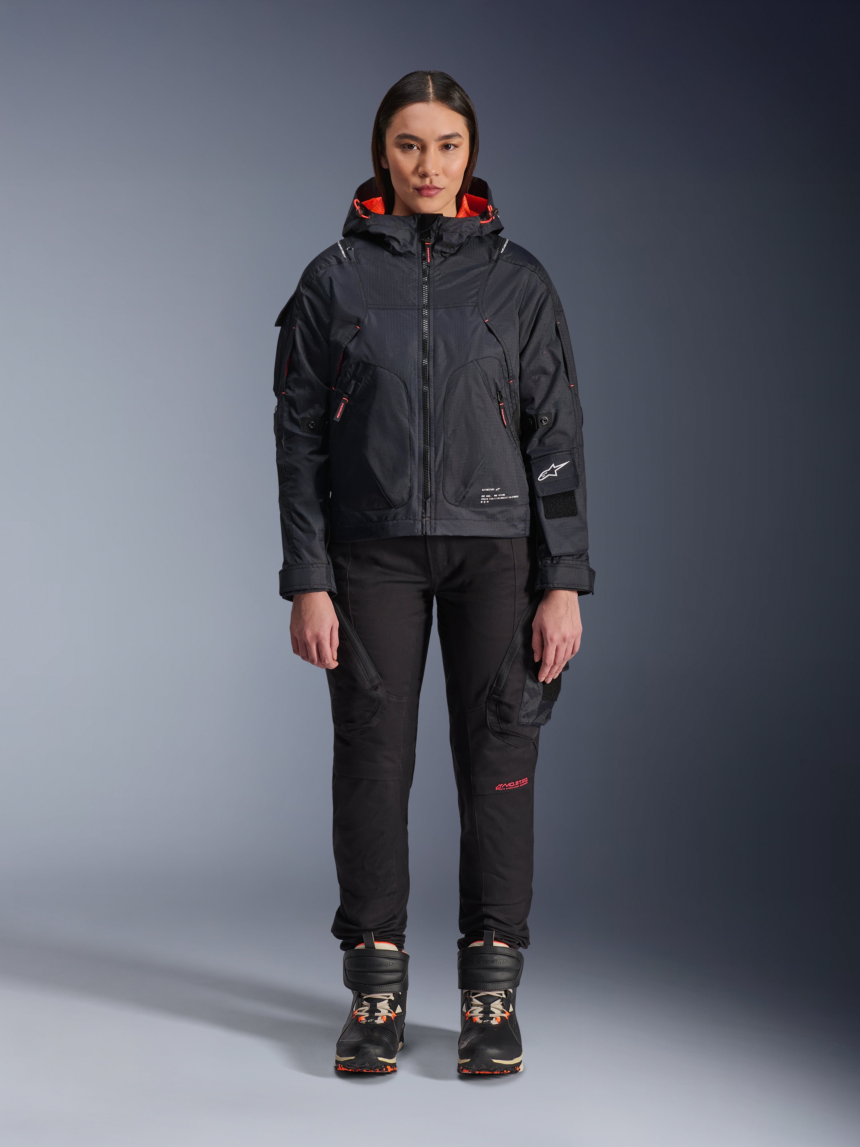 Vrouwelijk model draagt de Alpinestars Molly Air Jacket in Black en Fluo Coral, voorzien van een op utility geïnspireerd ontwerp met een capuchon en koraalrode binnenvoering, gecombineerd met een zwarte motorbroek en motorlaarzen, urban contemporary motoruitrusting