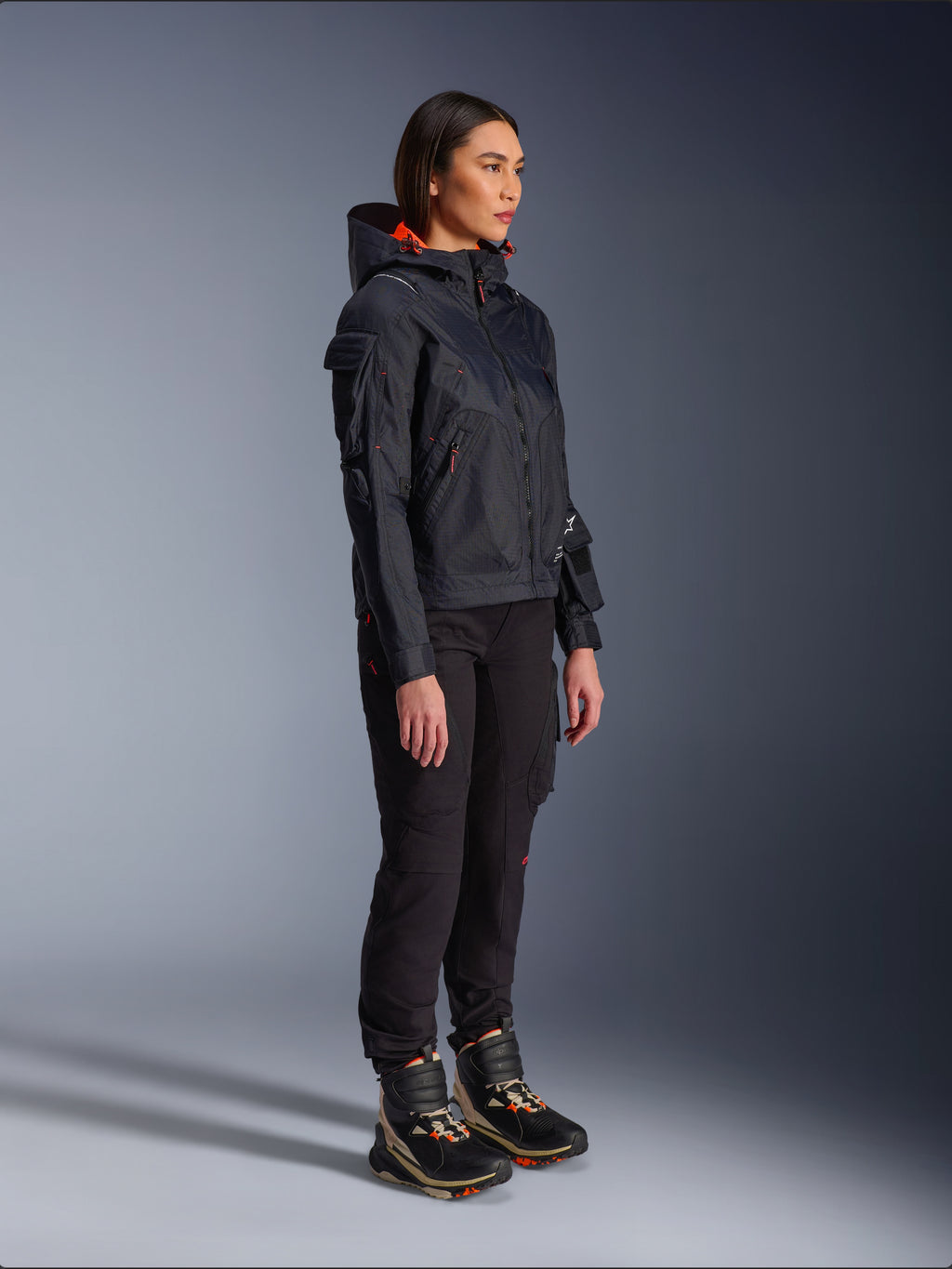Vrouwelijke motorrijder met de Alpinestars Molly Air motorjas in Black Fluo Coral, lichtgewicht textiele motorjas met een feloranje capuchonvoering, gecombineerd met een zwarte motorbroek en technische laarzen, urban contemporary utility-geïnspireerd ontwerp.