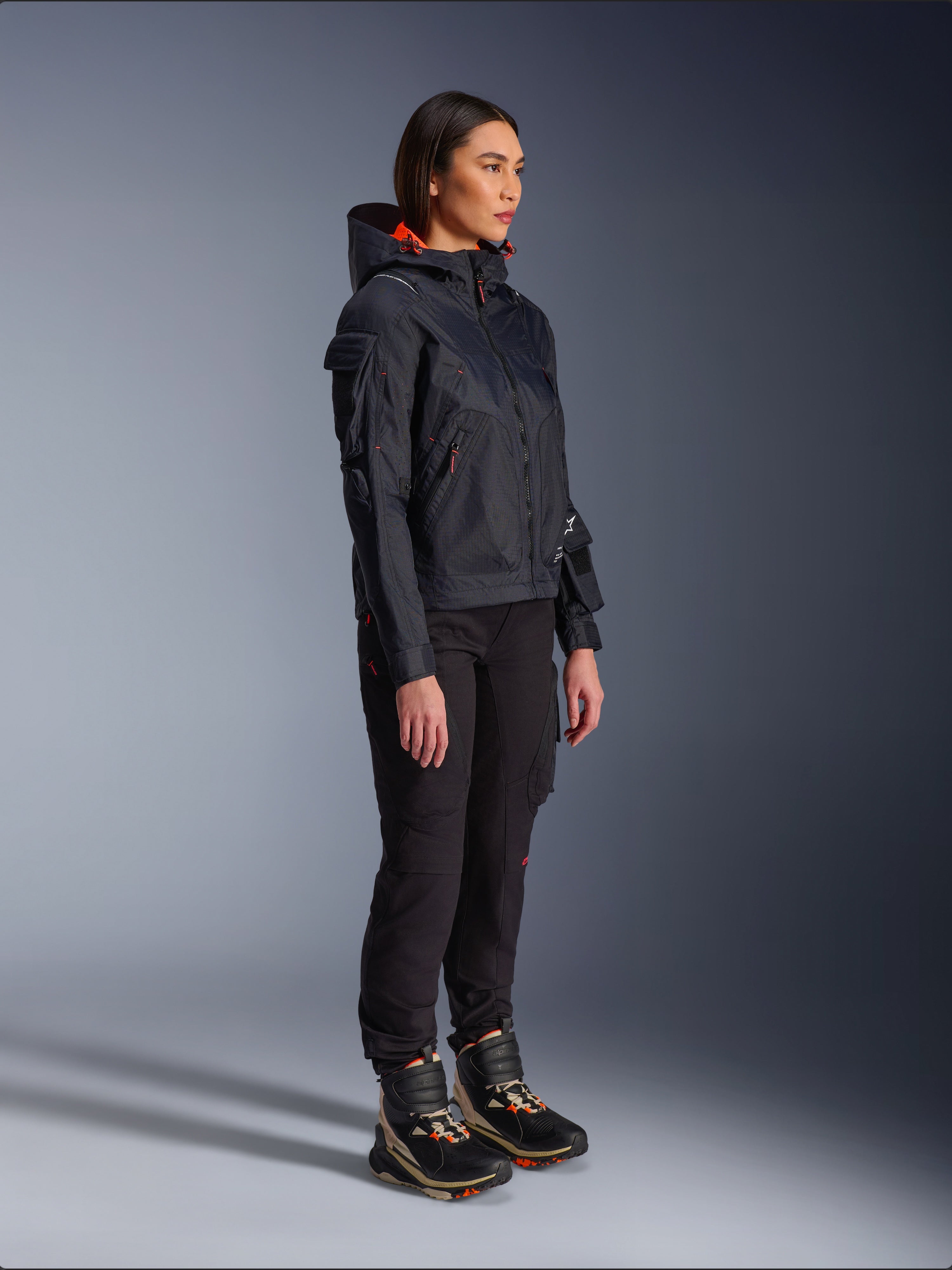 Vrouwelijke motorrijder met de Alpinestars Molly Air motorjas in Black Fluo Coral, lichtgewicht textiele motorjas met een feloranje capuchonvoering, gecombineerd met een zwarte motorbroek en technische laarzen, urban contemporary utility-geïnspireerd ontwerp.