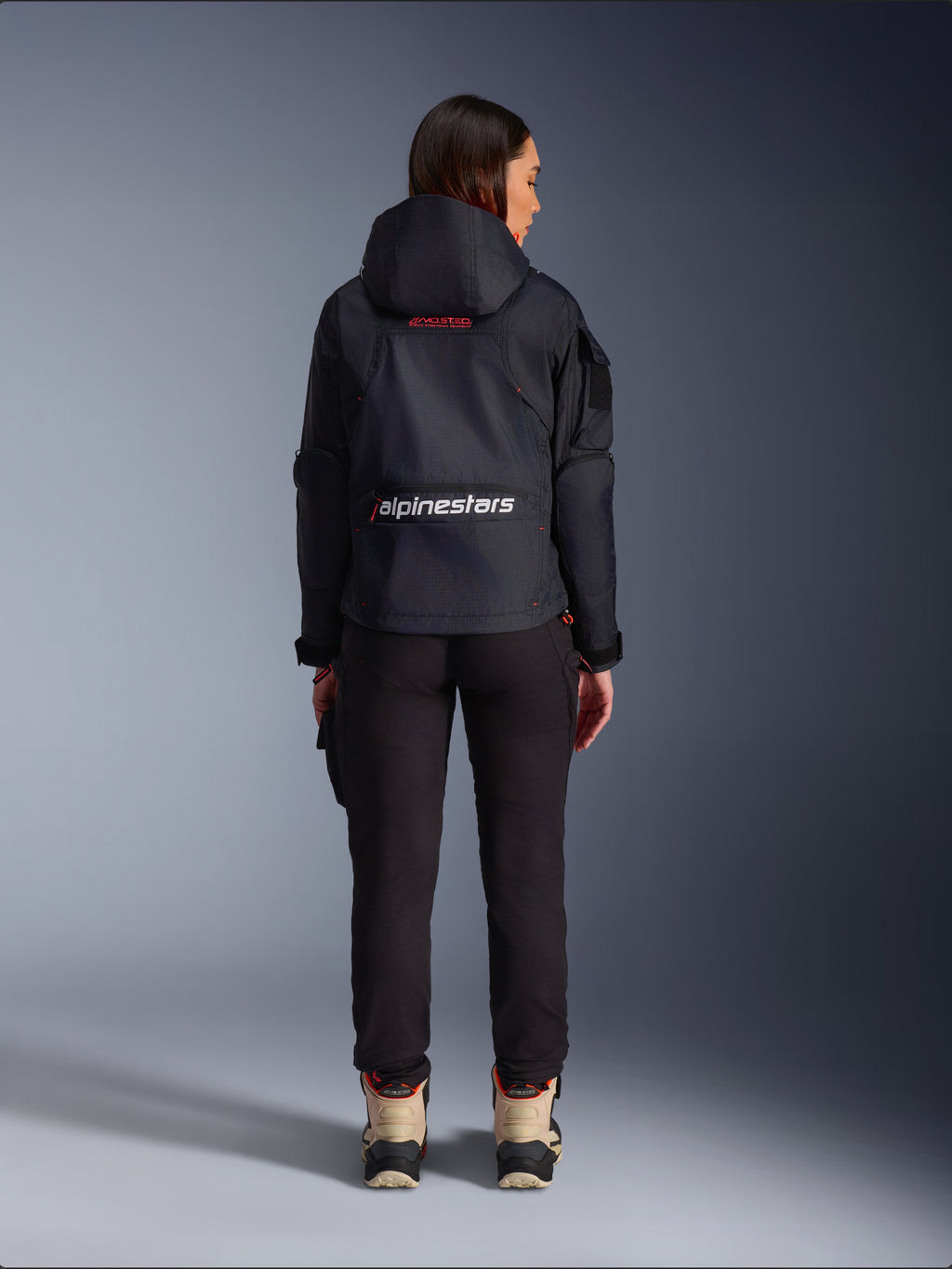 Alpinestars Actea damesbroek, zwarte motorbroek met een toelopende pasvorm, gecombineerd met een donkere Molly Air Jacket met capuchon en beige laarzen, achteraanzicht van een vrouwelijke rijder in urban motorkleding