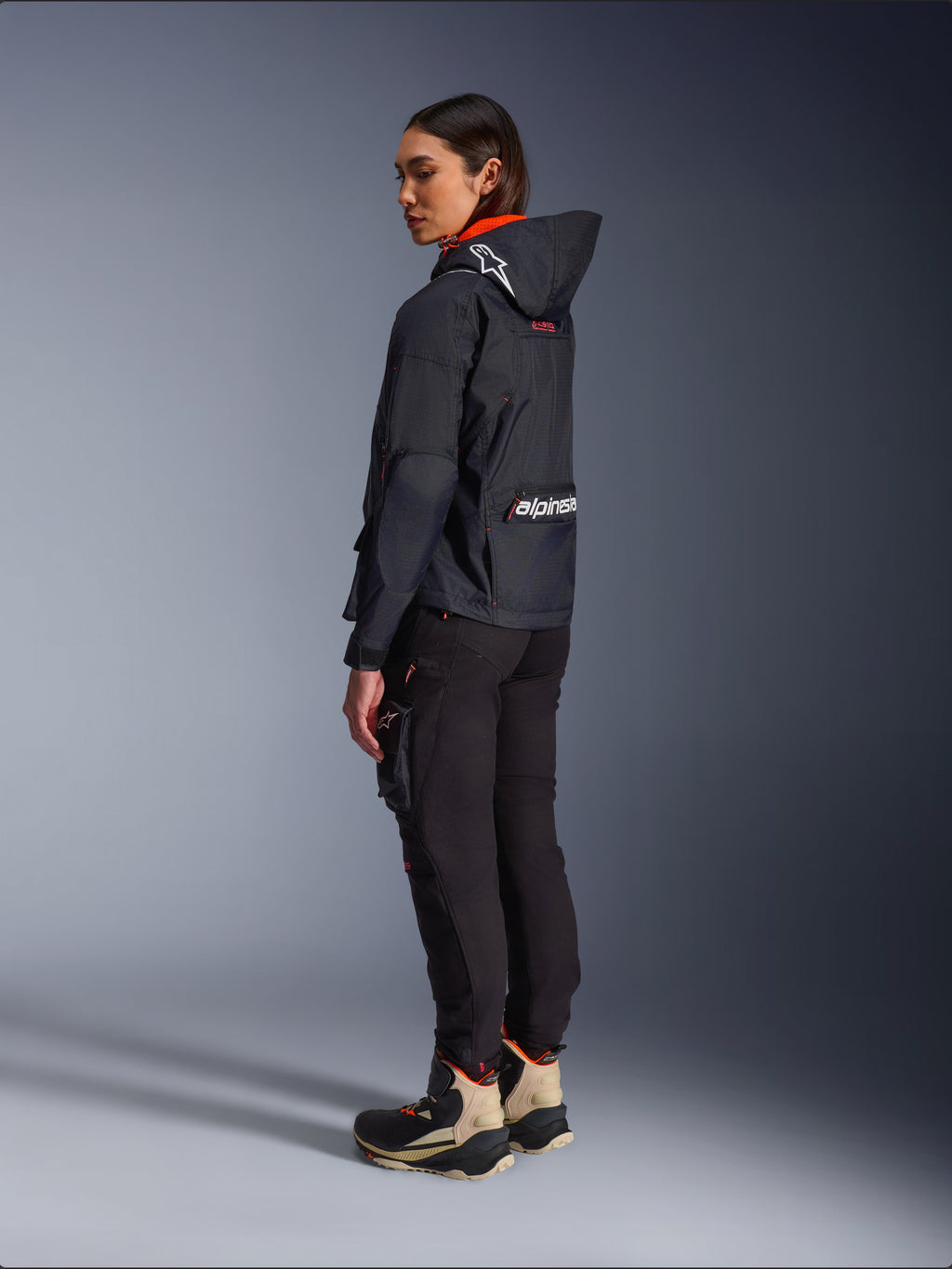 Alpinestars Actea dames motorbroek, zwart, moderne motorbroek met een taps toelopende pasvorm en cargo-zak op het bovenbeen, getoond vanuit een zij-achterperspectief op een model met een donker jack met capuchon en motorlaarzen, urban rijstijl.