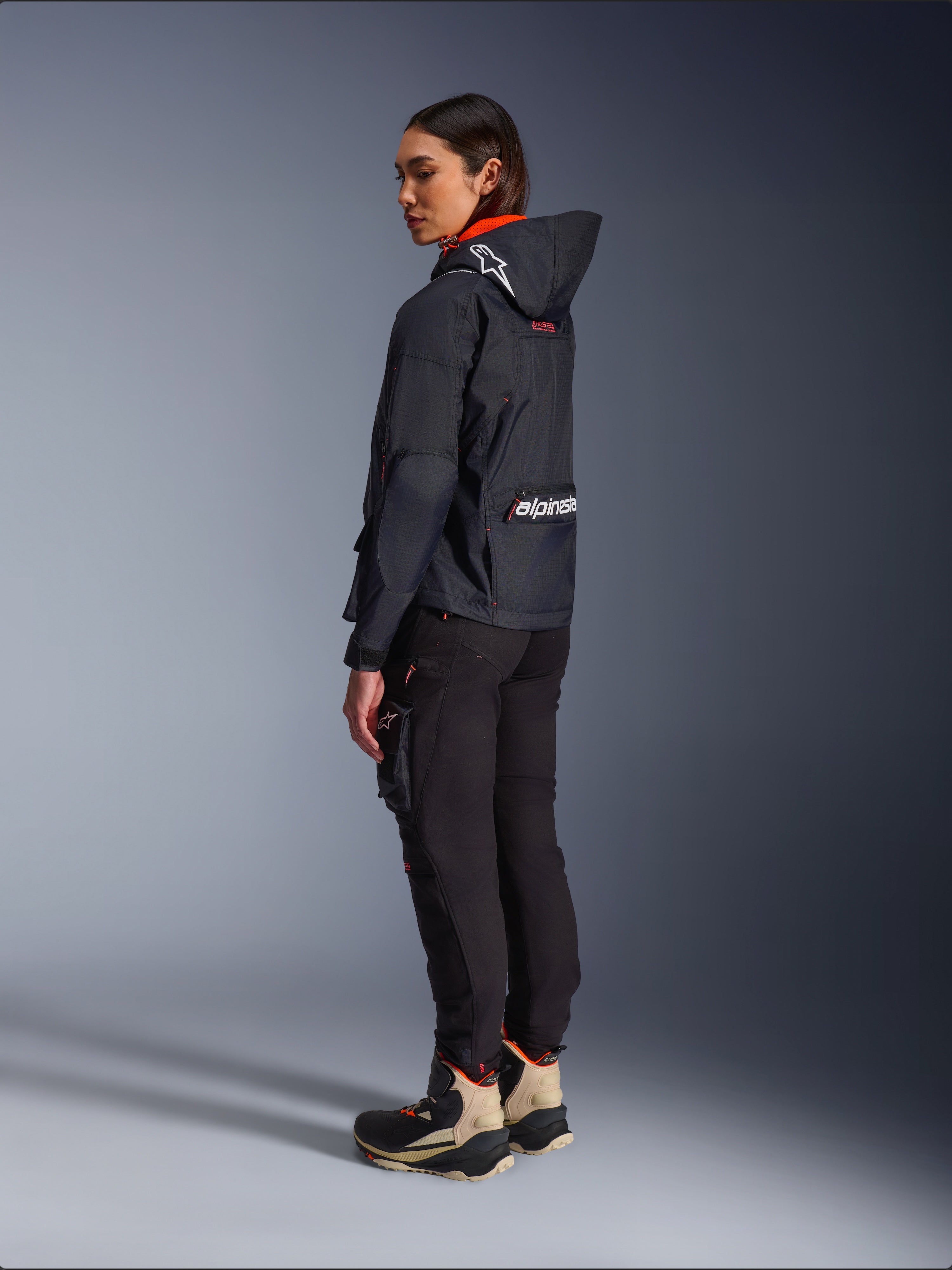 Alpinestars Actea dames motorbroek, zwart, moderne motorbroek met een taps toelopende pasvorm en cargo-zak op het bovenbeen, getoond vanuit een zij-achterperspectief op een model met een donker jack met capuchon en motorlaarzen, urban rijstijl.