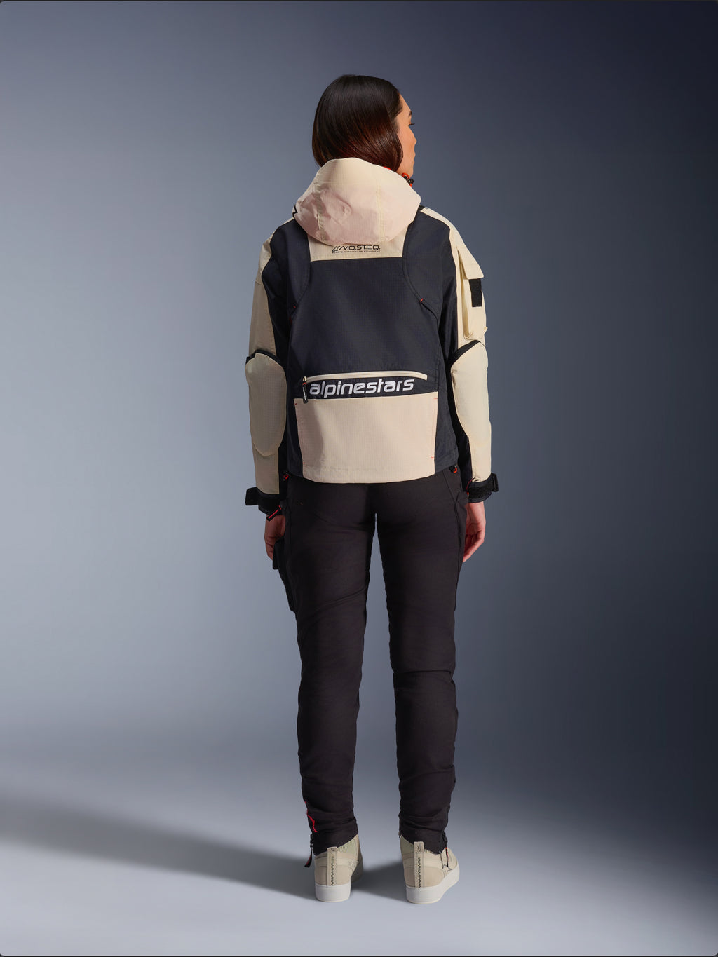 Alpinestars Molly Air Jacket, motorjas voor dames, Black Almond, achteraanzicht met de capuchon en groot logo op de rug, op utility geïnspireerd ripstop-ontwerp, gecombineerd met een zwarte motorbroek.