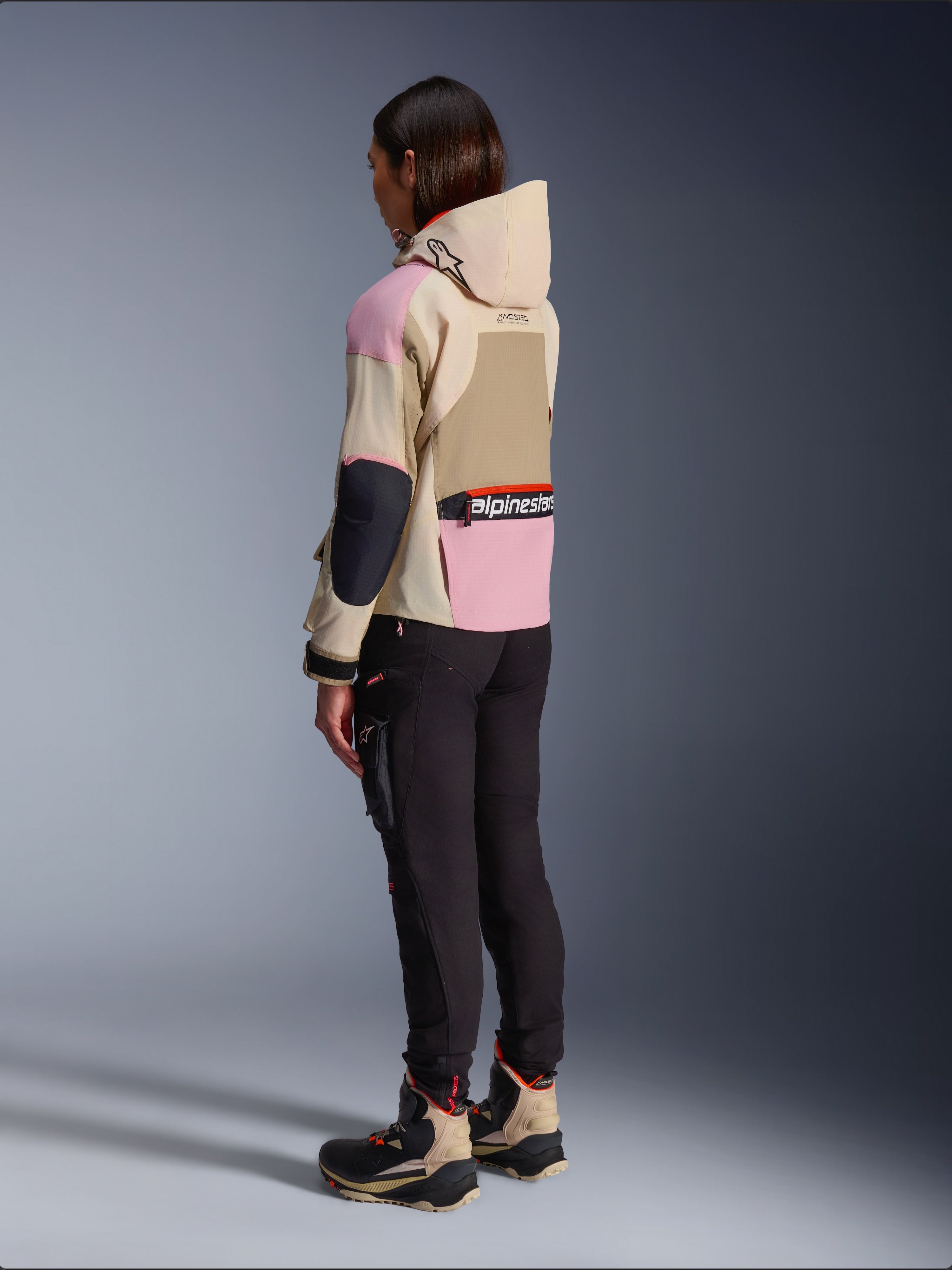 Female rider wearing the Alpinestars Molly Air Jacket in Sand Pink Almond, featuring a multi-tonal beige and pink design, shown from a rear side view with black riding pants and matching technical boots, urban motorcycle style with a hood.