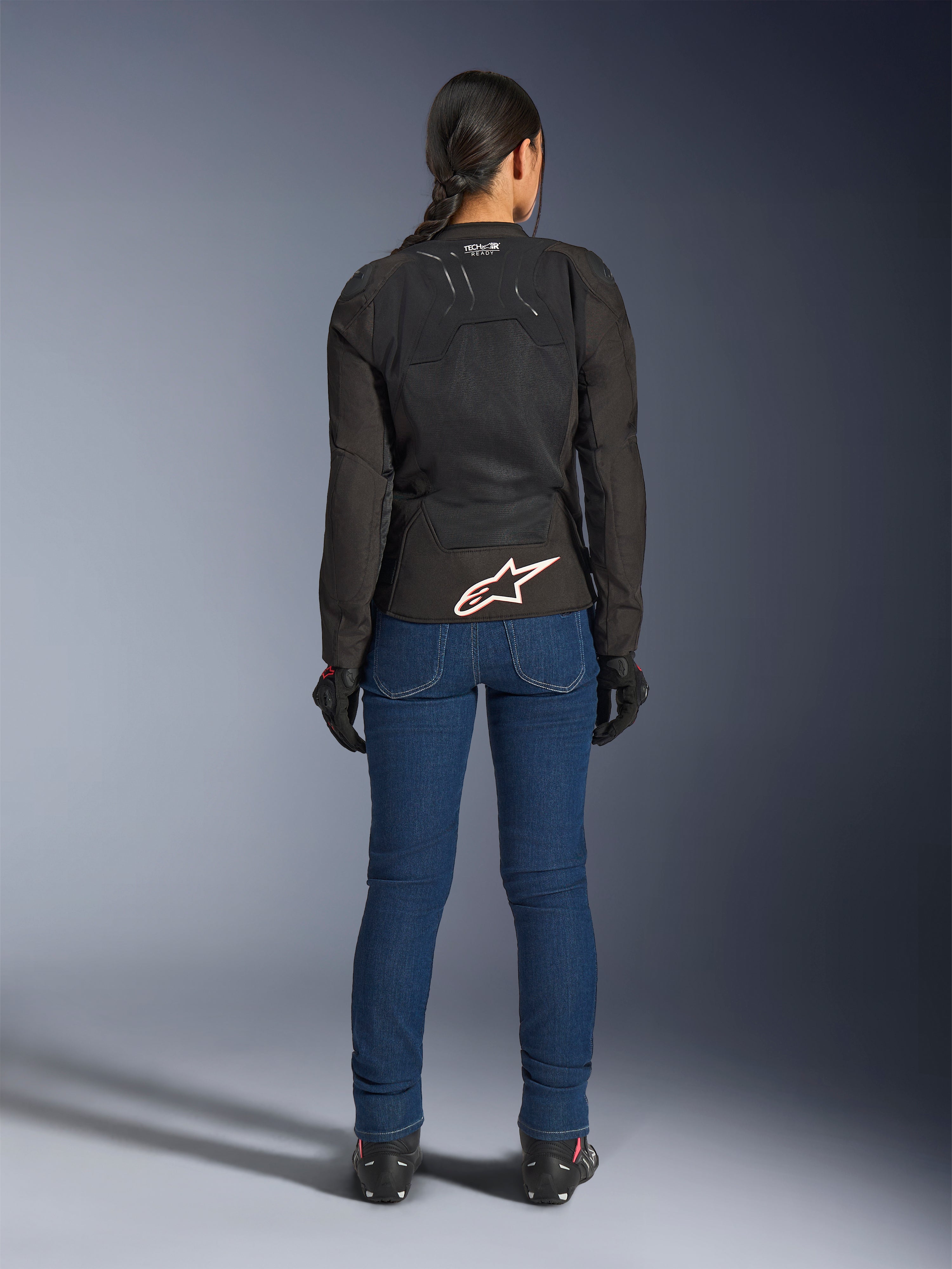 Vrouwelijke motorrijder van achteren gezien die de Alpinestars Stella SMX Air jas in het zwart draagt, gecombineerd met een blauwe denim motorbroek en zwarte handschoenen, voorzien van een geventileerd textielontwerp, een wit sterlogo op de onderrug en Tech-Air Ready-branding, ontworpen voor sportief rijden bij warm weer