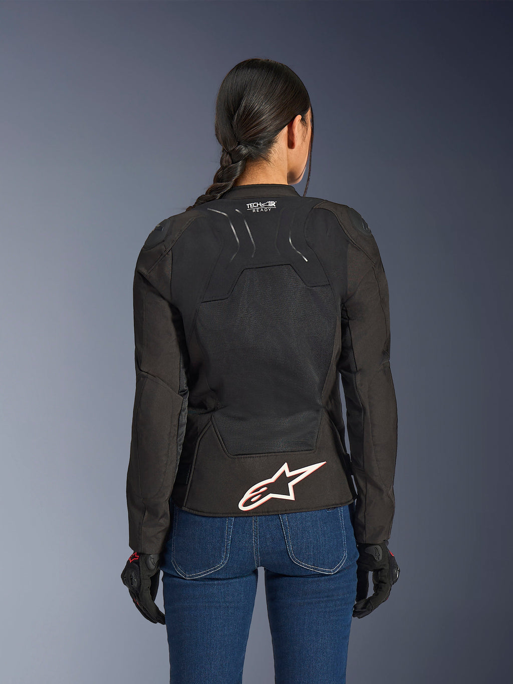 Alpinestars Stella SMX Air jas, achteraanzicht op vrouwelijk model, zwart textiel met high-flow mesh panelen, 'TECH-AIR READY' branding op de bovenrug, groot wit Alpinestars-logo met rode omtrek op de onderrug, gecombineerd met een blauwe spijkerbroek en zwarte motorhandschoenen
