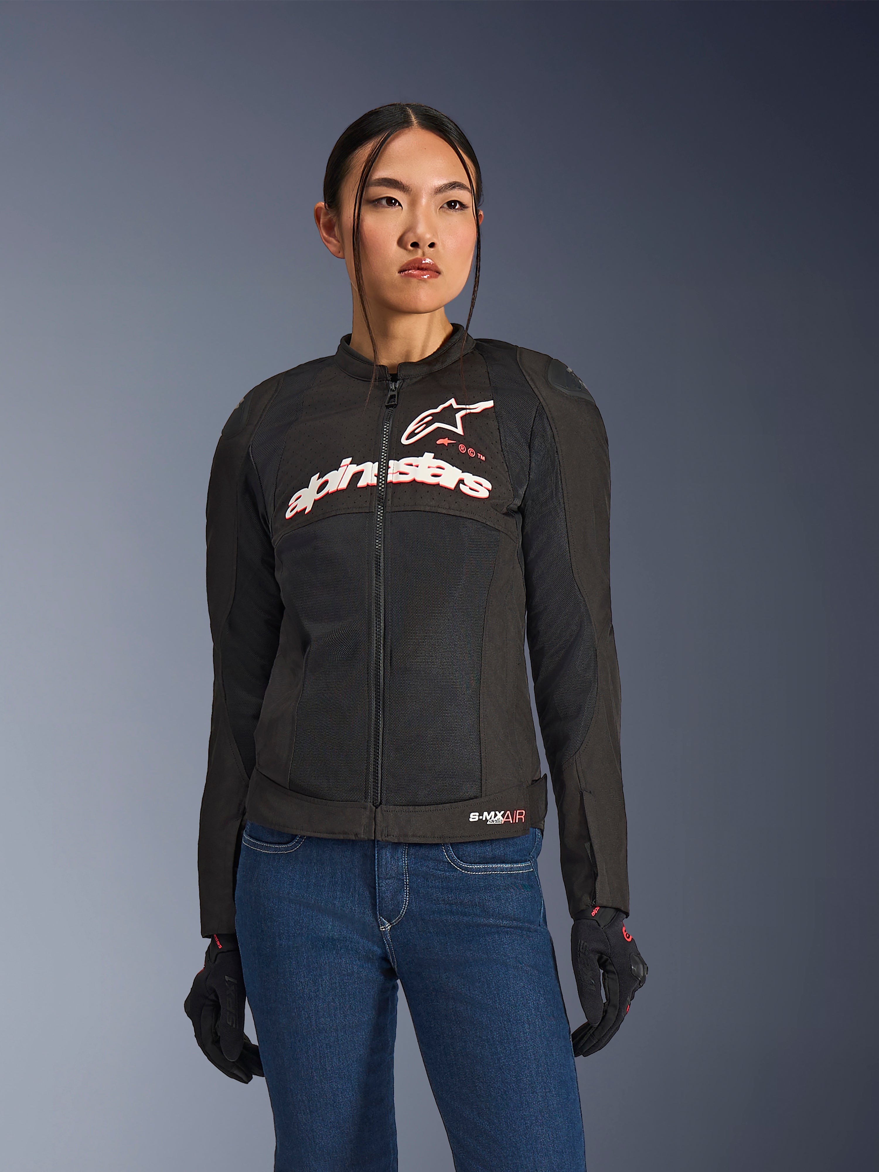 Vrouw draagt Alpinestars Stella SMX Air jas, Black White Red Fluo, textiele motorjas met geventileerd mesh en wit logo op de borst, gecombineerd met blauwe jeans en zwarte motorhandschoenen, vrouwelijke pasvorm voor sportief rijden