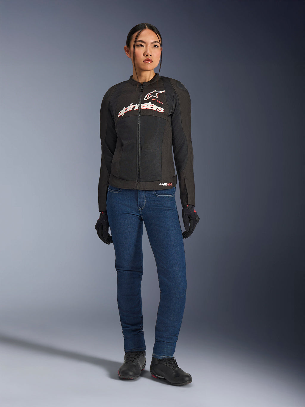 Vrouwelijke motorrijder draagt de Alpinestars Stella SMX Air jas, zwart met wit en Red Fluo logo's, gecombineerd met een blauwe denim motorbroek en zwarte handschoenen, sportieve geventileerde textieljas voor op de weg