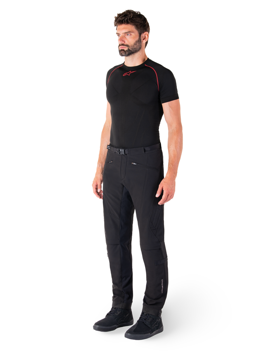 Alpinestars Alden broek, urban motorbroek, zwart, regular fit softshell textiel, gedragen door een model met een zwarte basislaag en rijschoenen, voorzien van een tailleband met magnetische sluiting en zakken met ritssluiting