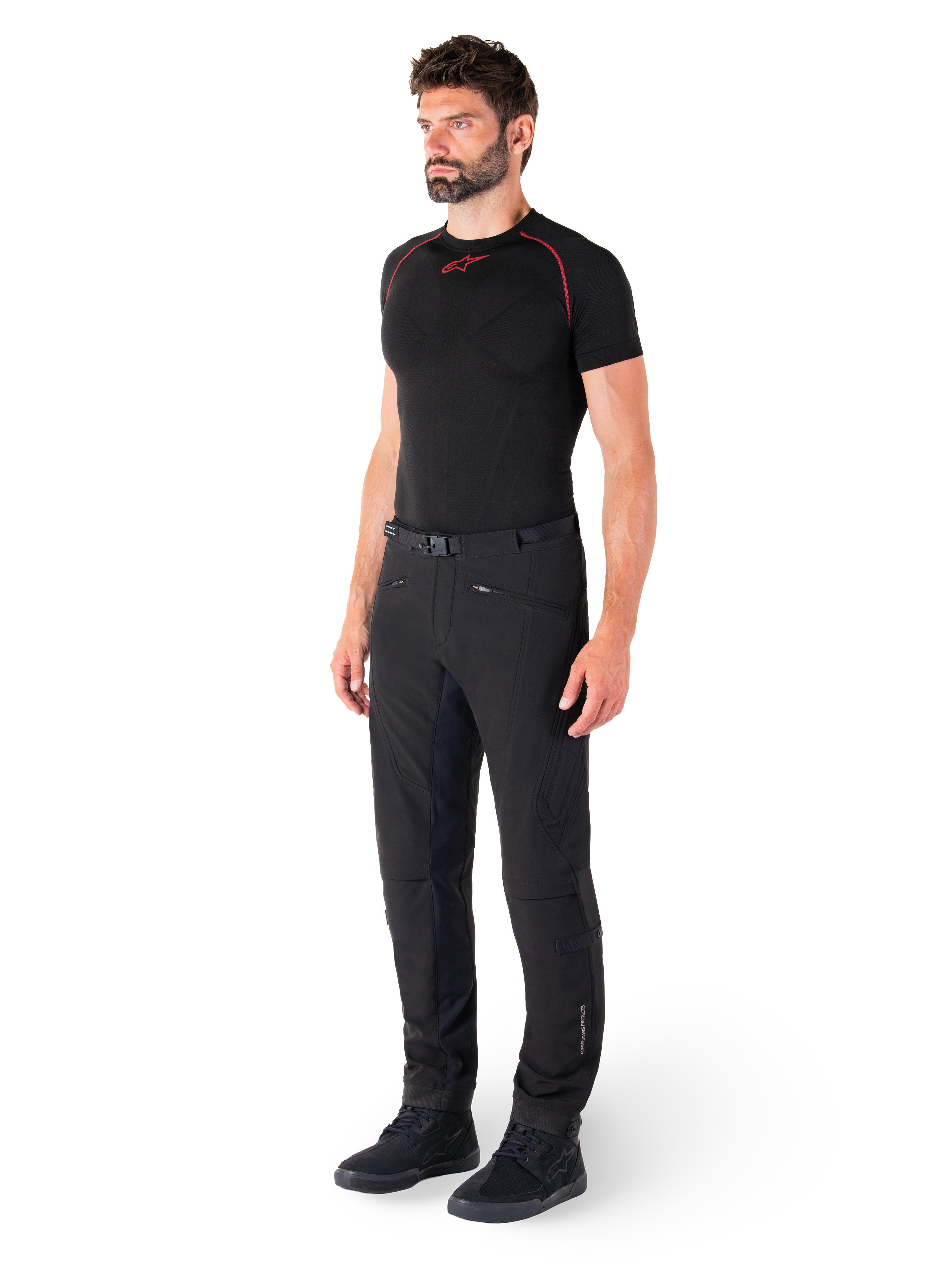 Alpinestars Alden broek, urban motorbroek, zwart, regular fit softshell textiel, gedragen door een model met een zwarte basislaag en rijschoenen, voorzien van een tailleband met magnetische sluiting en zakken met ritssluiting