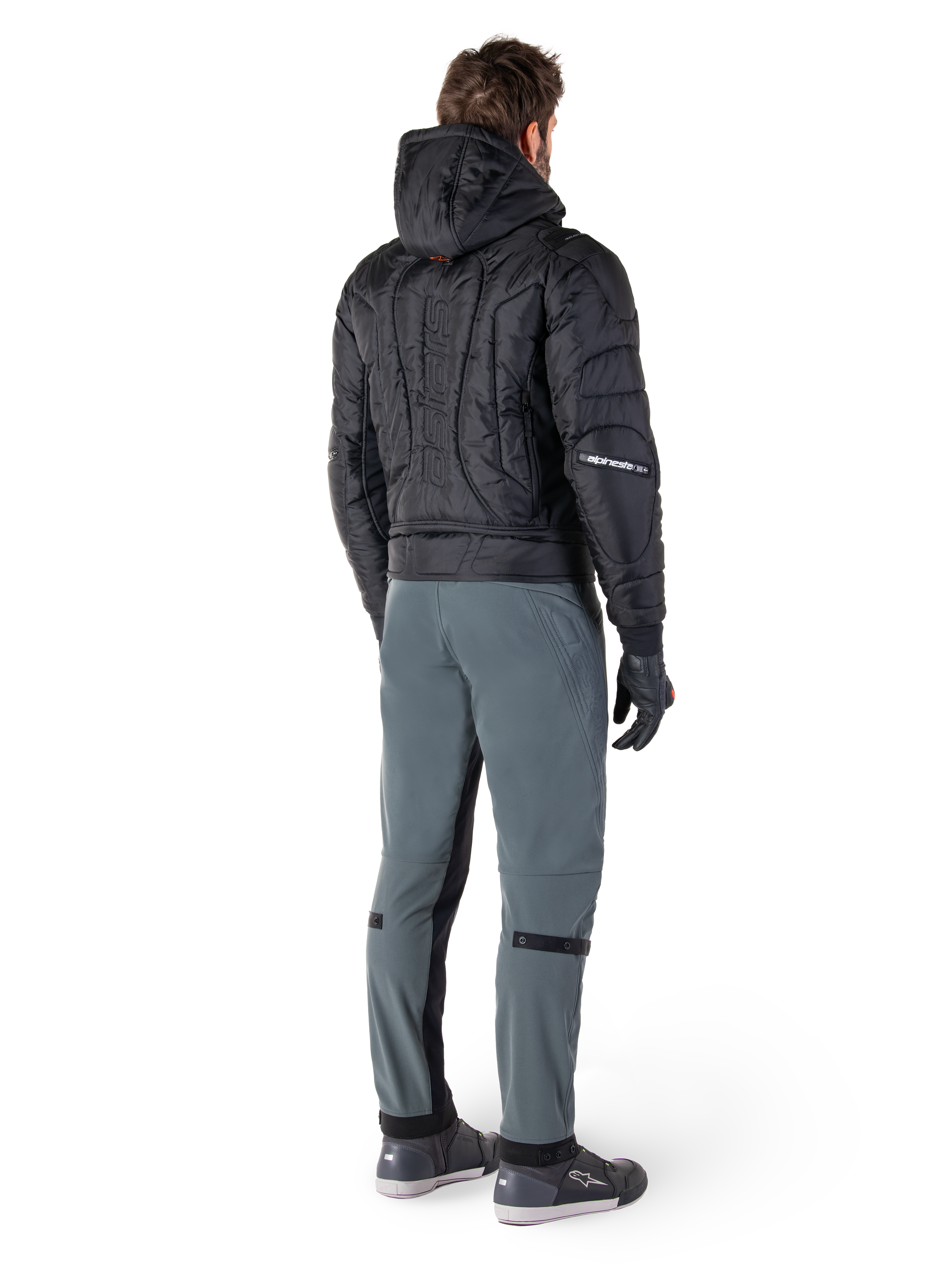 Alpinestars Alden broek, urban motorbroek, Urban Gray, achteraanzicht van een motorrijder die een zwart gewatteerd jack met capuchon en motorsneakers draagt, voorzien van een slanke pasvorm onder de knie met zwarte stretchpanelen en knieverstellers, modern op streetwear geïnspireerd ontwerp.