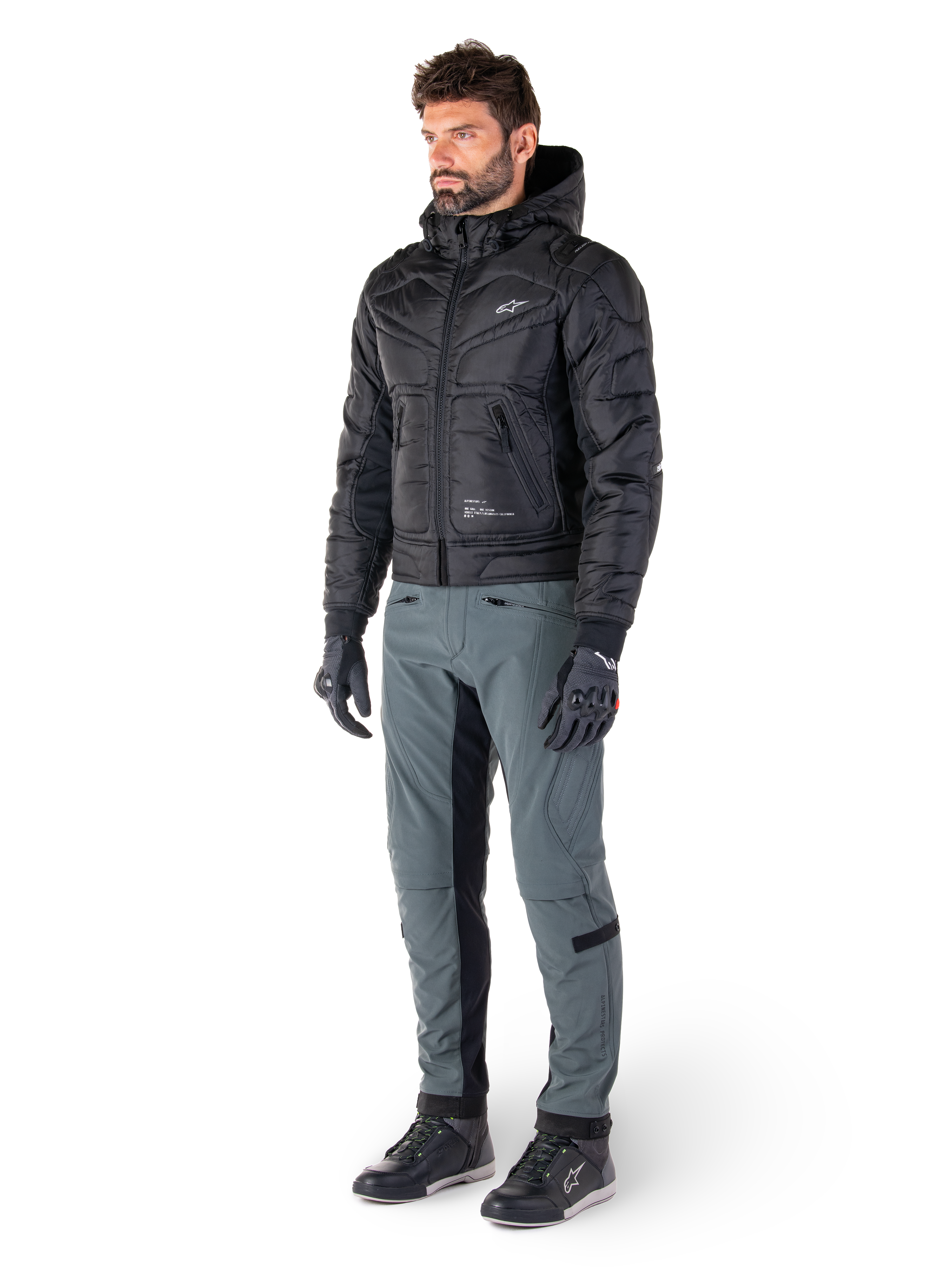 Alpinestars Alden broek in Urban Gray, gedragen door een model met een zwart gewatteerd jack met capuchon, handschoenen en rijschoenen, voorzien van gewatteerde details aan de zijkanten en zwarte binnenbeenpanelen, streetwear-geïnspireerde stedelijke motoruitrusting.