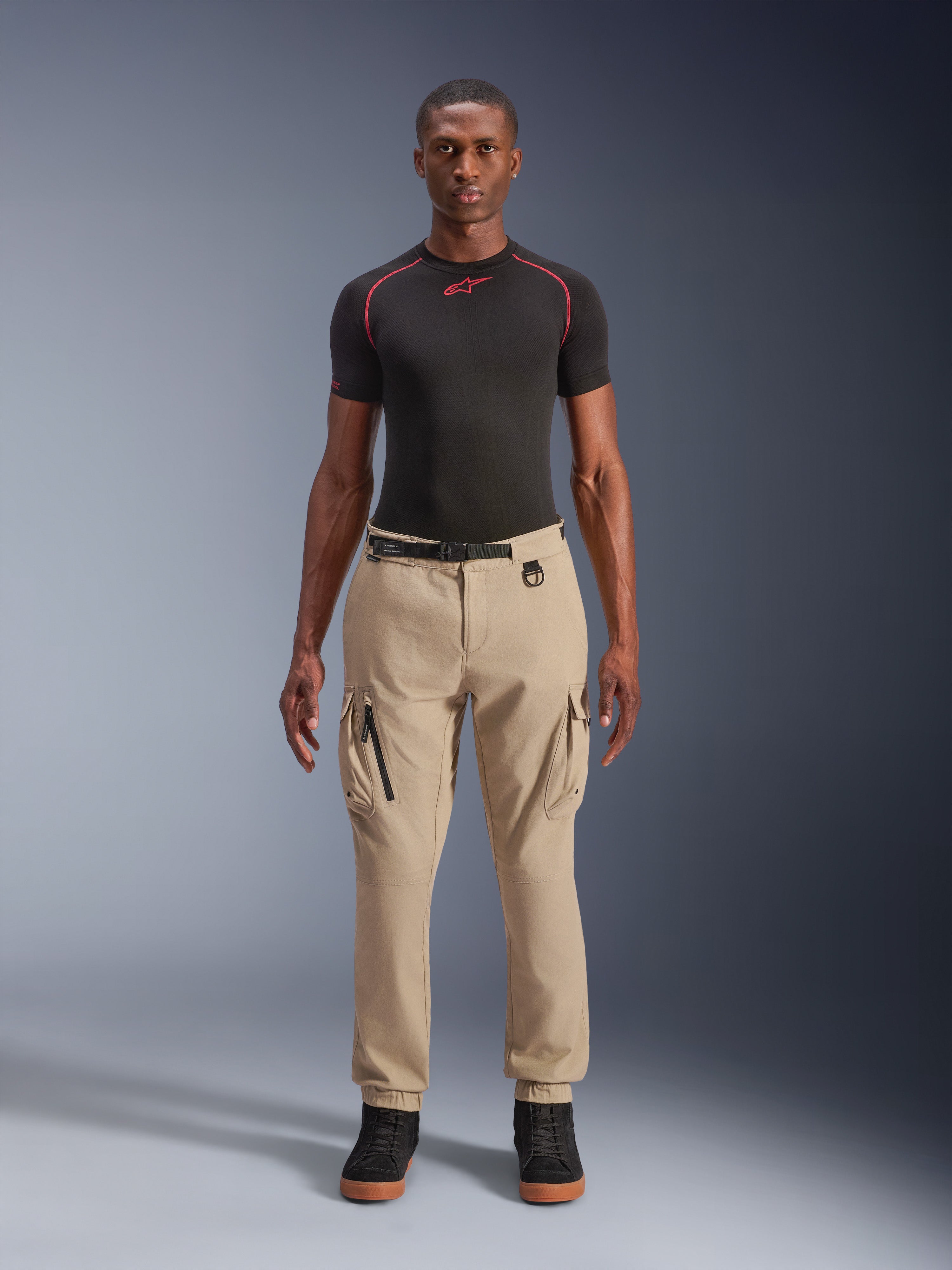 Alpinestars Flex-AST Cargo Pants - Short, lichtbeige urban motorbroek, regular tapered pasvorm met elastische zomen, functionele cargozakken en geïntegreerde zwarte riem met gesp, gedragen door een mannelijk model met een zwarte basislaag en sneakers.