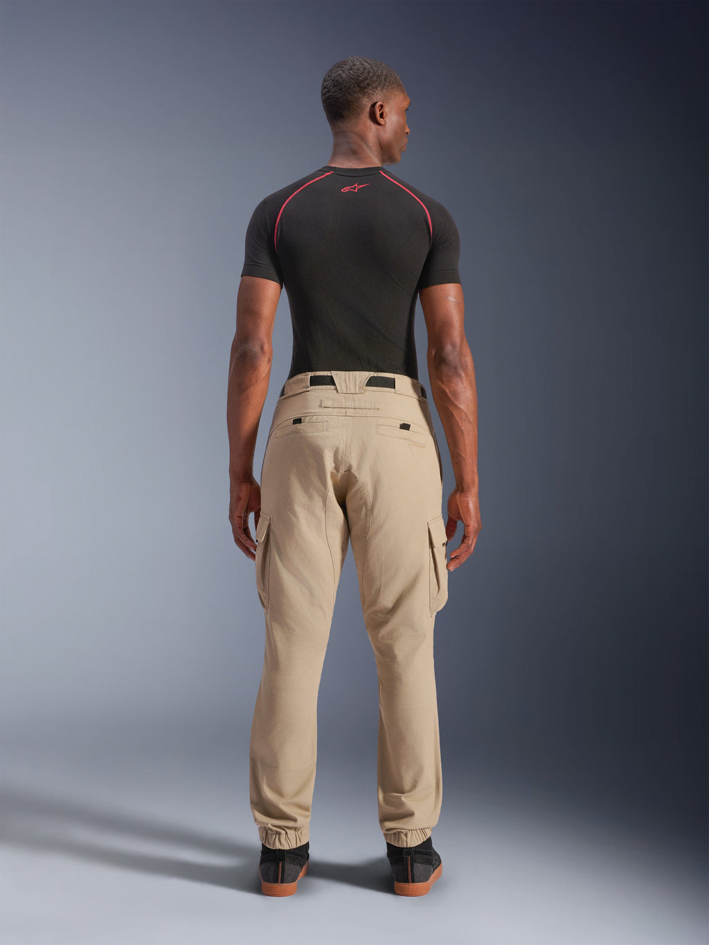 Alpinestars Flex-AST Cargo Broek - Short, lichtbeige, achteraanzicht op een model, regular tapered pasvorm met elastische zomen en cargozakken, gedragen met een zwarte technische basislaag met rode accenten, moderne urban motorkleding
