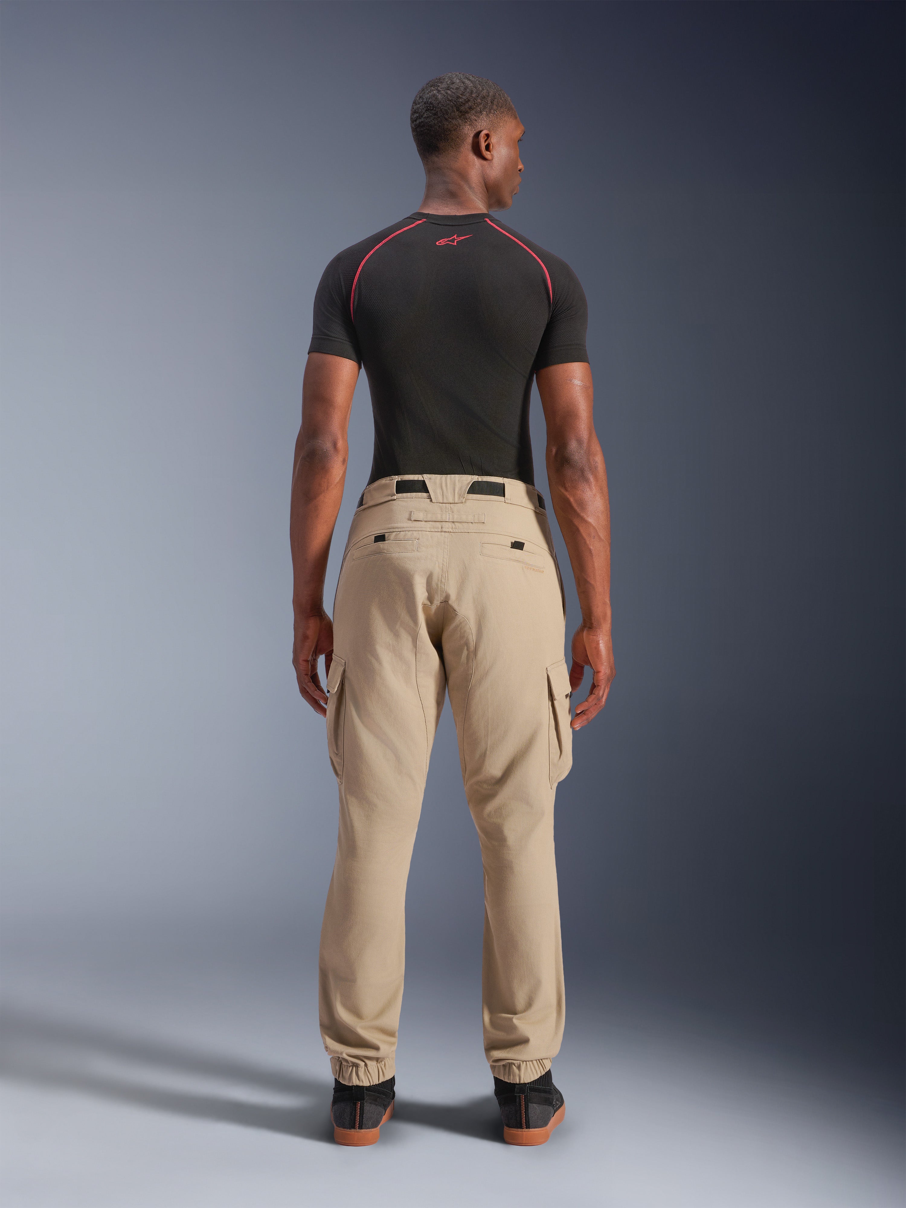 Alpinestars Flex-AST Cargo Broek - Short, lichtbeige, achteraanzicht op een model, regular tapered pasvorm met elastische zomen en cargozakken, gedragen met een zwarte technische basislaag met rode accenten, moderne urban motorkleding