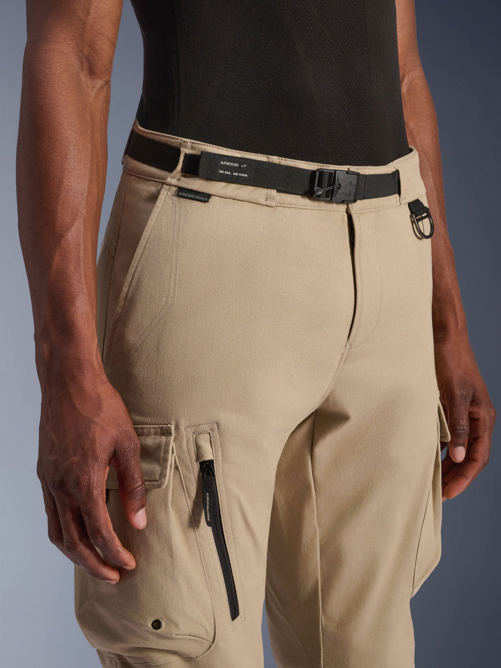 Alpinestars Flex-AST Cargo broek - Kort, lichtbeige, close-up van de taille en het bovenbeen, met een zwarte verstelbare riem met Alpinestars-branding, metalen D-ring en functionele cargozakken met ritsen en kleppen met drukknoppen, urban contemporary motorkleding