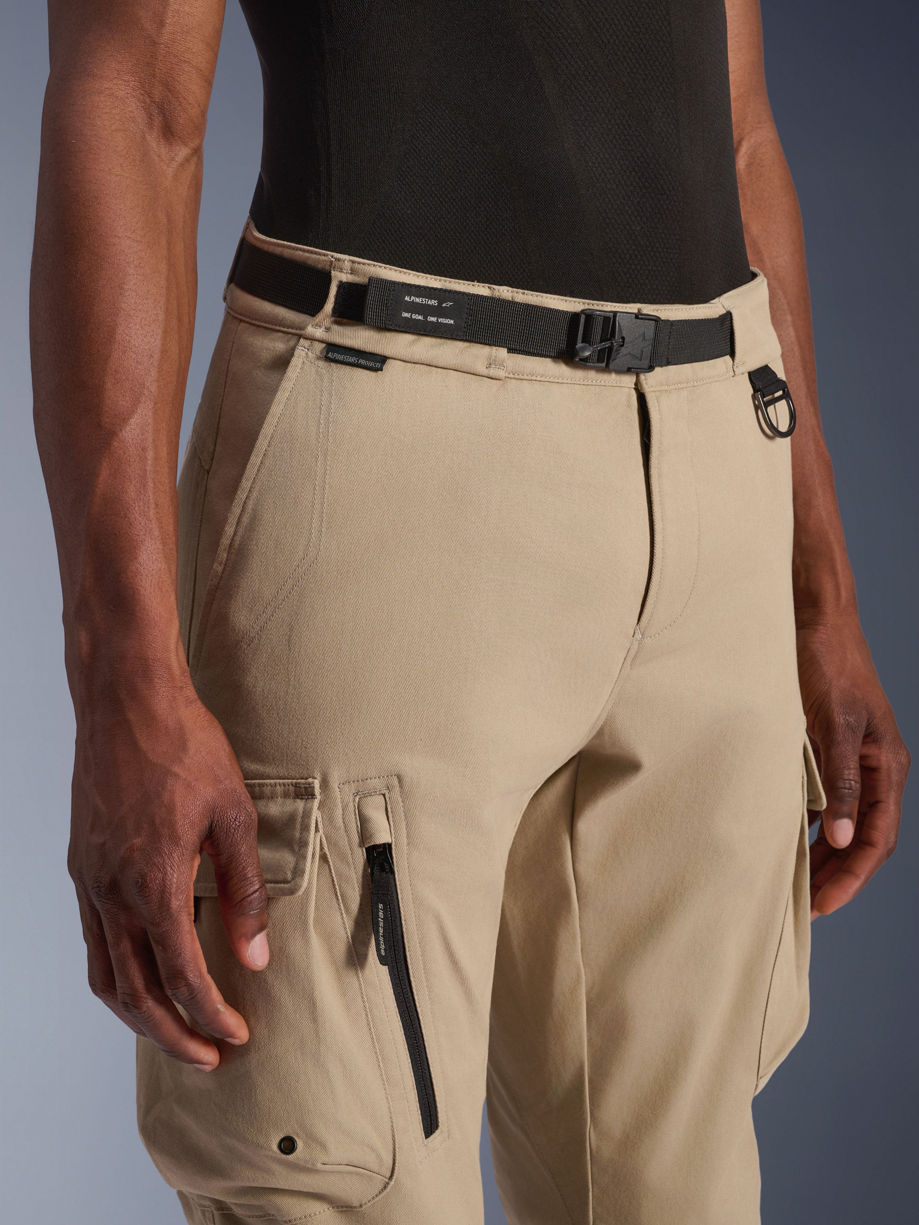 Alpinestars Flex-AST Cargo broek - Kort, lichtbeige, close-up van de taille en het bovenbeen, met een zwarte verstelbare riem met Alpinestars-branding, metalen D-ring en functionele cargozakken met ritsen en kleppen met drukknoppen, urban contemporary motorkleding