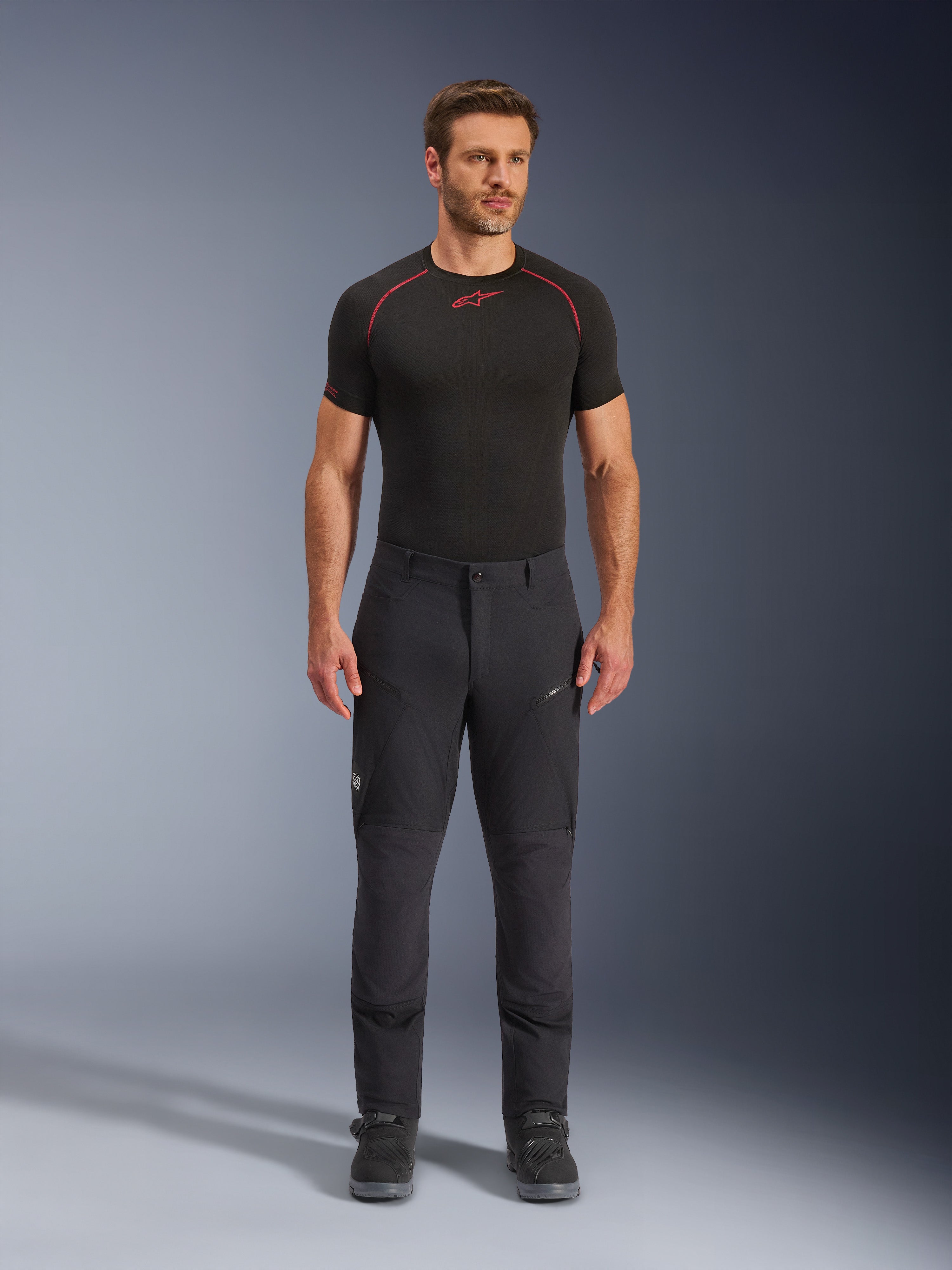 Alpinestars Flex-AST Explorer Pants, zwarte adventure touring motorbroek, man draagt nauwsluitende textiele rijbroek met cargozakken met rits en verstevigde panelen, gecombineerd met een zwarte basislaag en motorlaarzen, duurzame bescherming voor adventure rijden