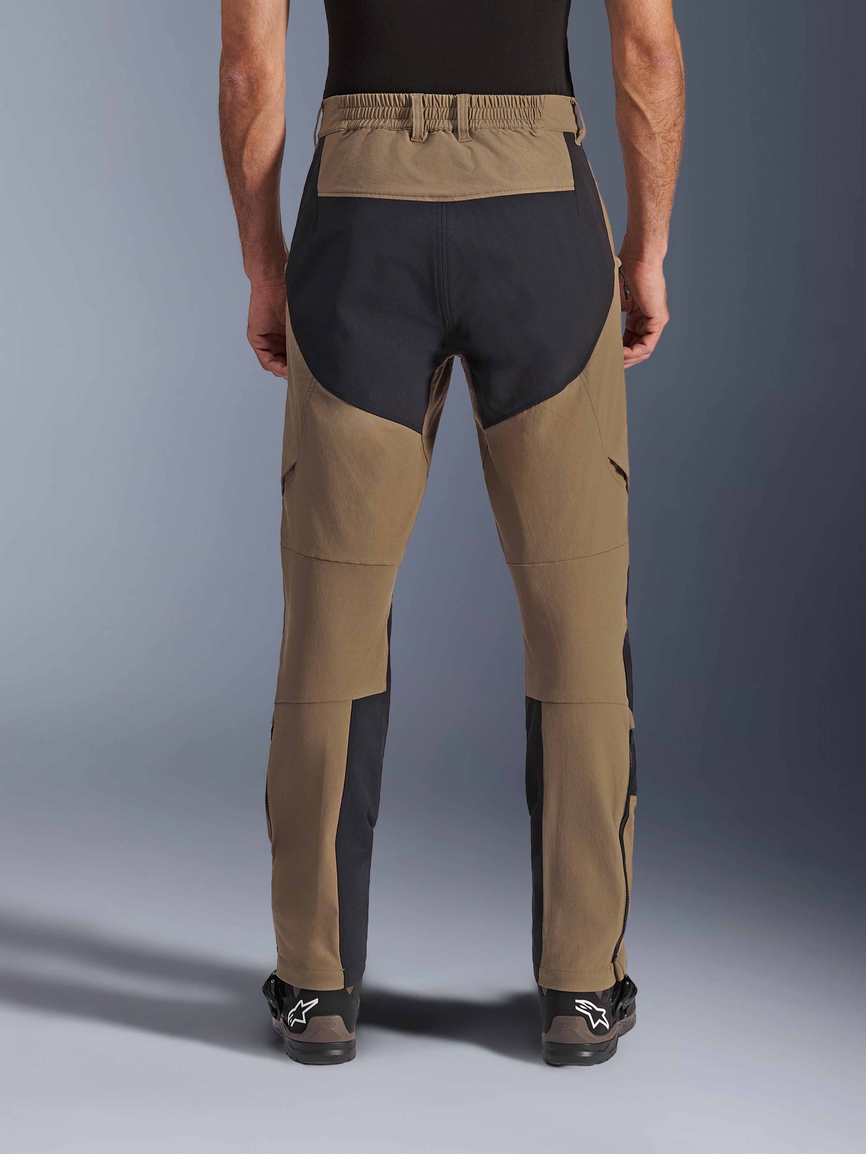 Alpinestars Flex-AST Explorer Pants, adventure motorbroek, Walnut Black, achteraanzicht gedragen door rijder, zwarte ripstop-verstevigingspanelen op het zitvlak en de binnenkant van de benen, elastische tailleband, lange ritsen aan het onderbeen en bijpassende adventure-laarzen