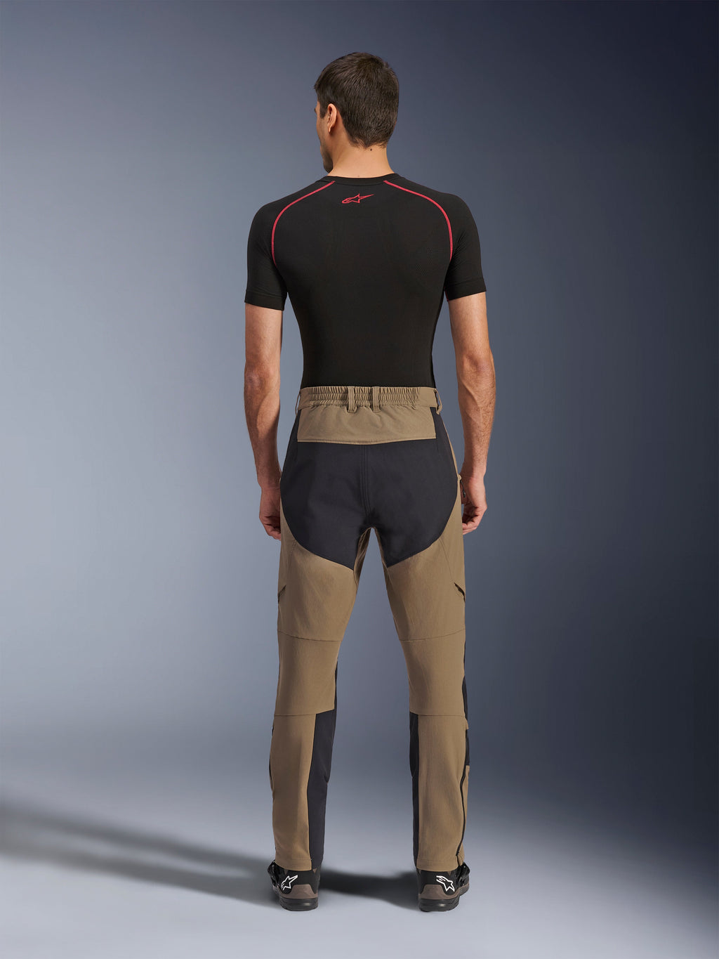 Alpinestars Flex-AST Explorer broek, Walnut Black, achteraanzicht op model, met versterkt zwart zitvlak en bruin stretchtextiel, gecombineerd met een zwarte base layer en technische motorlaarzen, duurzame adventure motorkleding