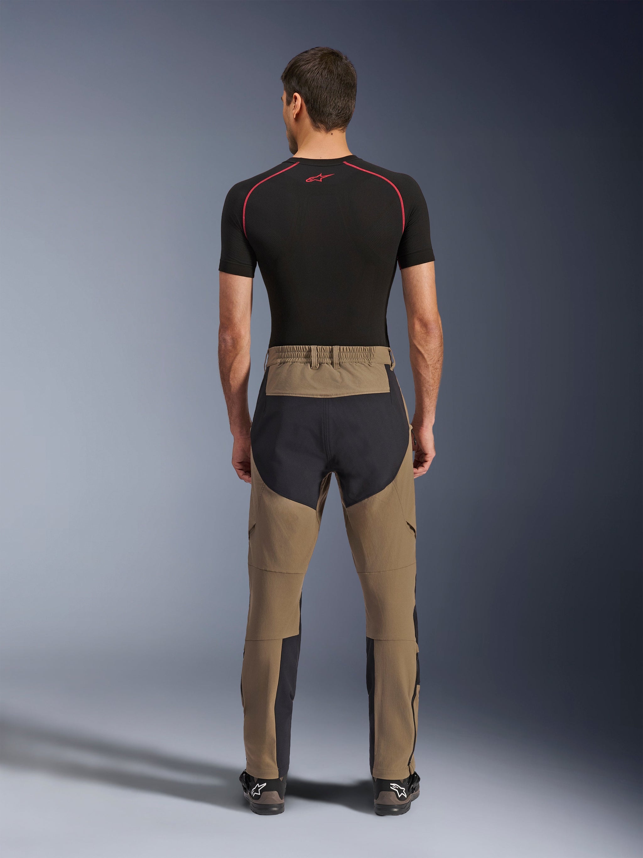 Alpinestars Flex-AST Explorer broek, Walnut Black, achteraanzicht op model, met versterkt zwart zitvlak en bruin stretchtextiel, gecombineerd met een zwarte base layer en technische motorlaarzen, duurzame adventure motorkleding