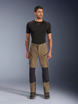 Alpinestars Flex-AST Explorer broek, adventure motorbroek, Walnut Black, gedragen door een model met een zwarte basislaag en laarzen, voorzien van beige stretchtextiel met zwarte versterkte kniepanelen en cargozakken met ritssluiting, ontworpen voor adventure touring