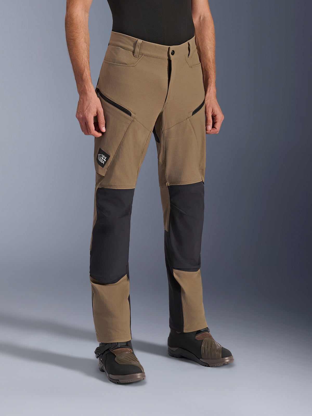 Alpinestars Flex-AST Explorer broek, adventure motorbroek in Walnut Black, voorzien van tan textiel met zwarte verstevigde knie- en zitvlakpanelen, cargozakken met ritssluiting en All Terra-logopatch, gedragen met technische motorlaarzen.