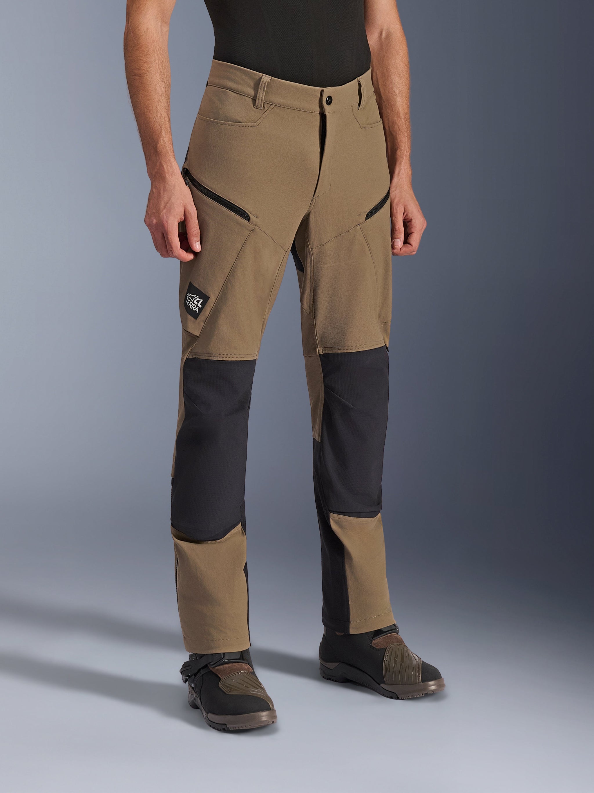 Alpinestars Flex-AST Explorer broek, adventure motorbroek in Walnut Black, voorzien van tan textiel met zwarte verstevigde knie- en zitvlakpanelen, cargozakken met ritssluiting en All Terra-logopatch, gedragen met technische motorlaarzen.