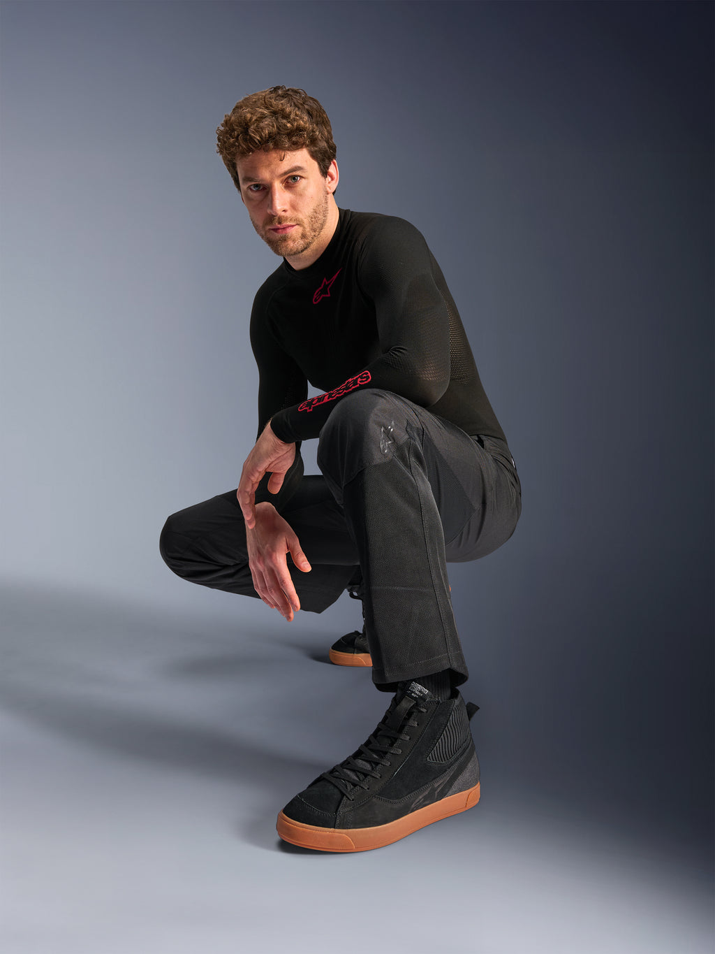 Alpinestars C-1 Air broek, urban motorbroek, zwart, model hurkend in een studio-setting, gedragen met een zwarte top met lange mouwen en zwarte sneakers met bruine zolen, voorzien van ademende mesh-panelen en een eigentijdse urban stijl