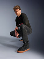 Alpinestars C-1 Air broek, urban motorbroek, zwart, model hurkend in een studio-setting, gedragen met een zwarte top met lange mouwen en zwarte sneakers met bruine zolen, voorzien van ademende mesh-panelen en een eigentijdse urban stijl