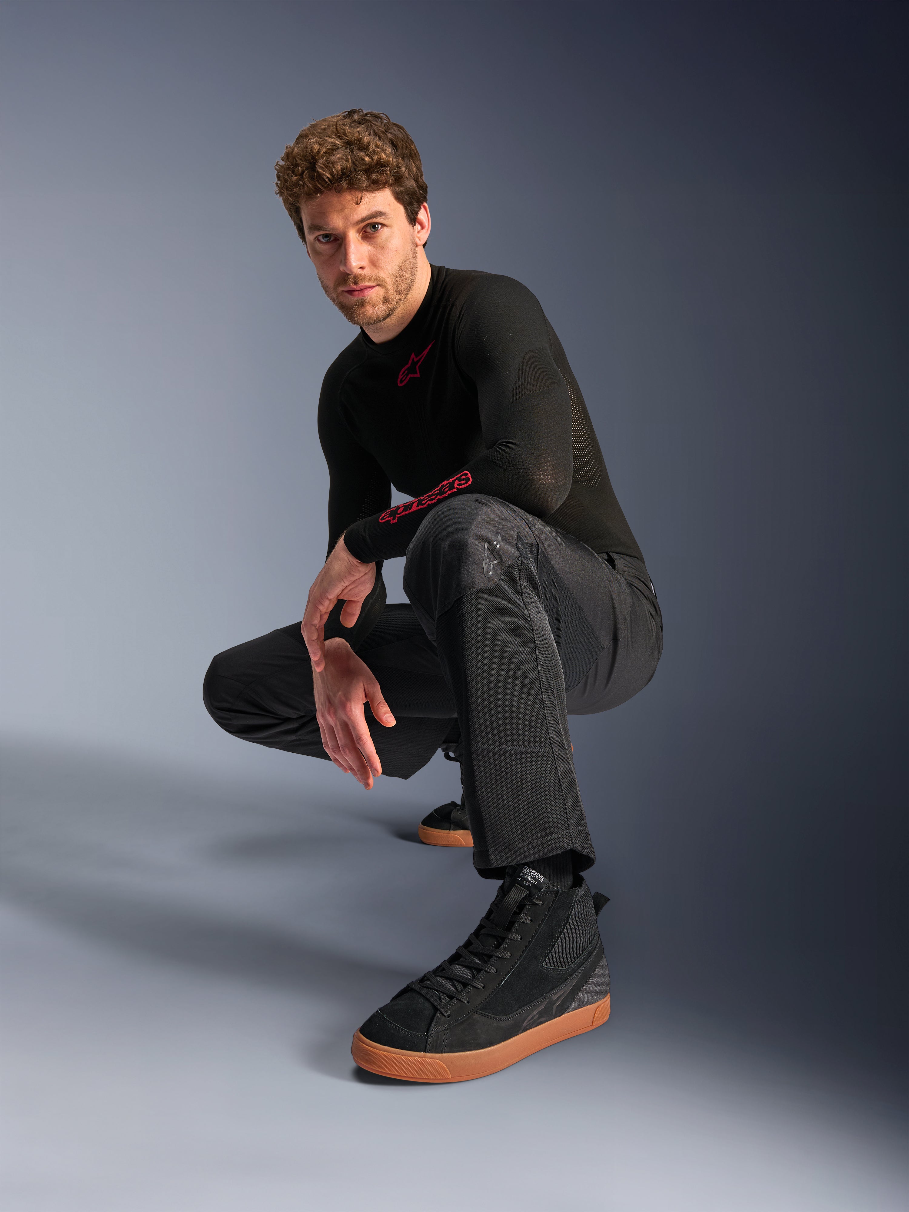 Alpinestars C-1 Air broek, urban motorbroek, zwart, model hurkend in een studio-setting, gedragen met een zwarte top met lange mouwen en zwarte sneakers met bruine zolen, voorzien van ademende mesh-panelen en een eigentijdse urban stijl