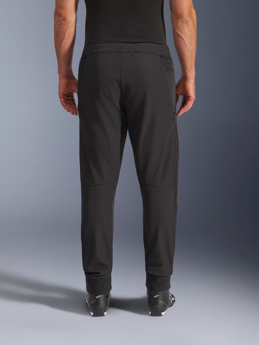 Alpinestars Aeroshell Airflow broek, zwart, achteraanzicht, met lasergeperforeerde zones op de kuiten en mesh-panelen, gedragen met zwarte Alpinestars laarzen, lichtgewicht softshell-ontwerp voor sport en touring