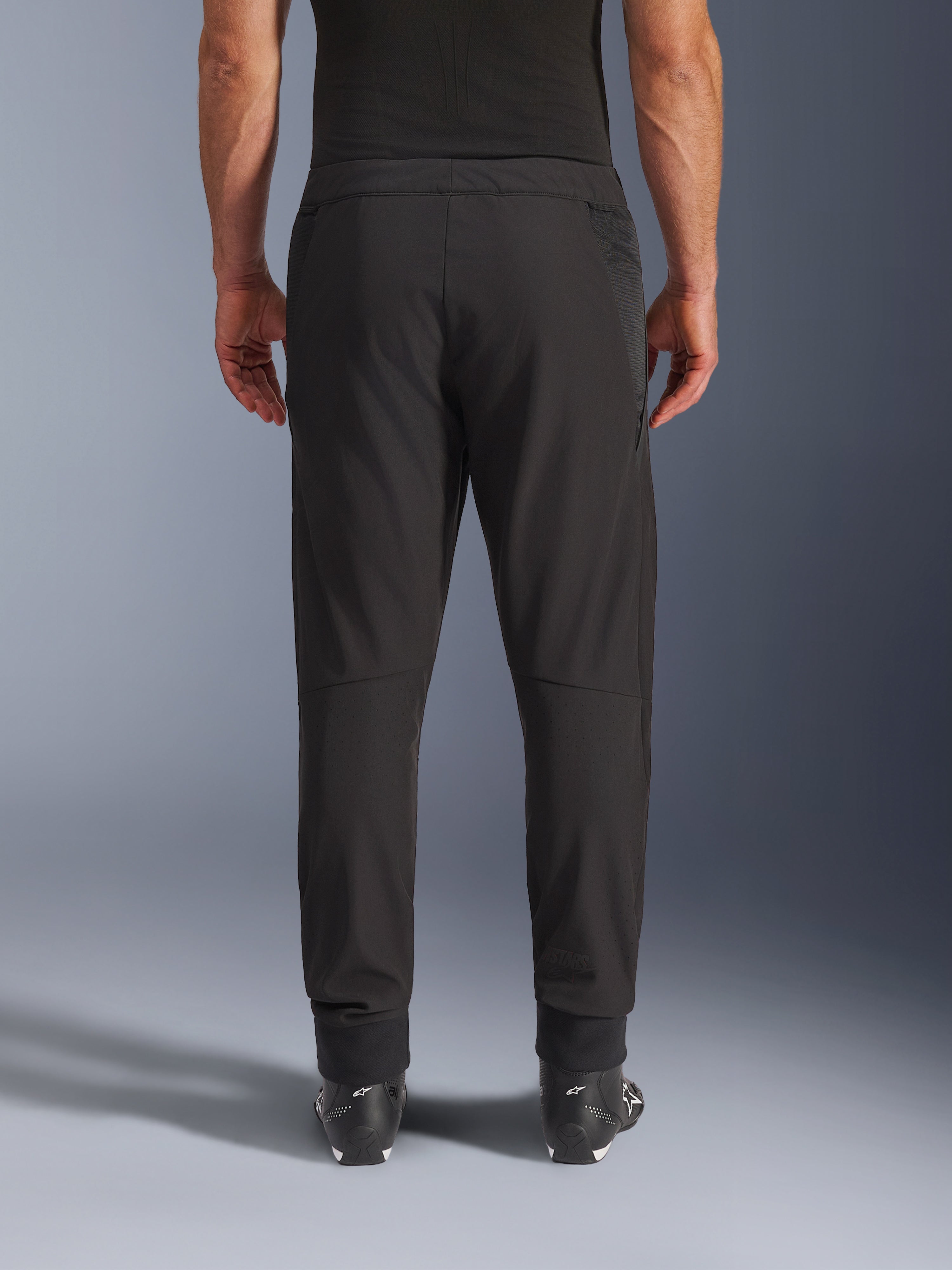 Alpinestars Aeroshell Airflow broek, zwart, achteraanzicht, met lasergeperforeerde zones op de kuiten en mesh-panelen, gedragen met zwarte Alpinestars laarzen, lichtgewicht softshell-ontwerp voor sport en touring