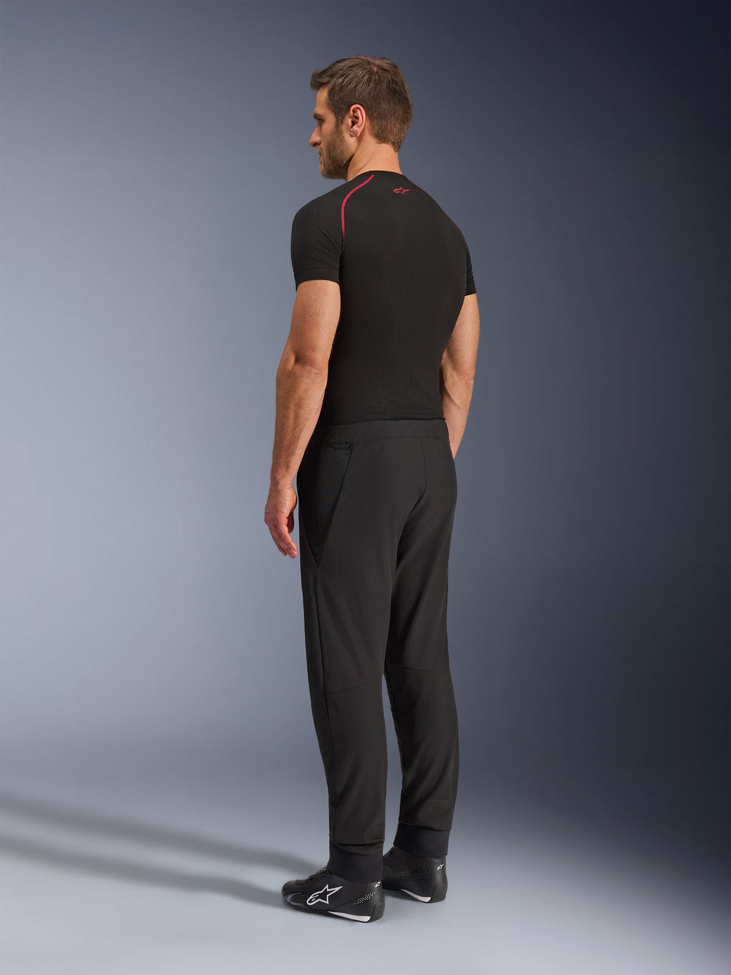 Alpinestars Aeroshell Airflow broek, zwarte motorbroek, lichtgewicht softshell textiel, gedragen met een zwart technisch shirt met korte mouwen en zwarte motorlaarzen, achteraanzicht in profiel toont de slanke pasvorm en ergonomische constructie.