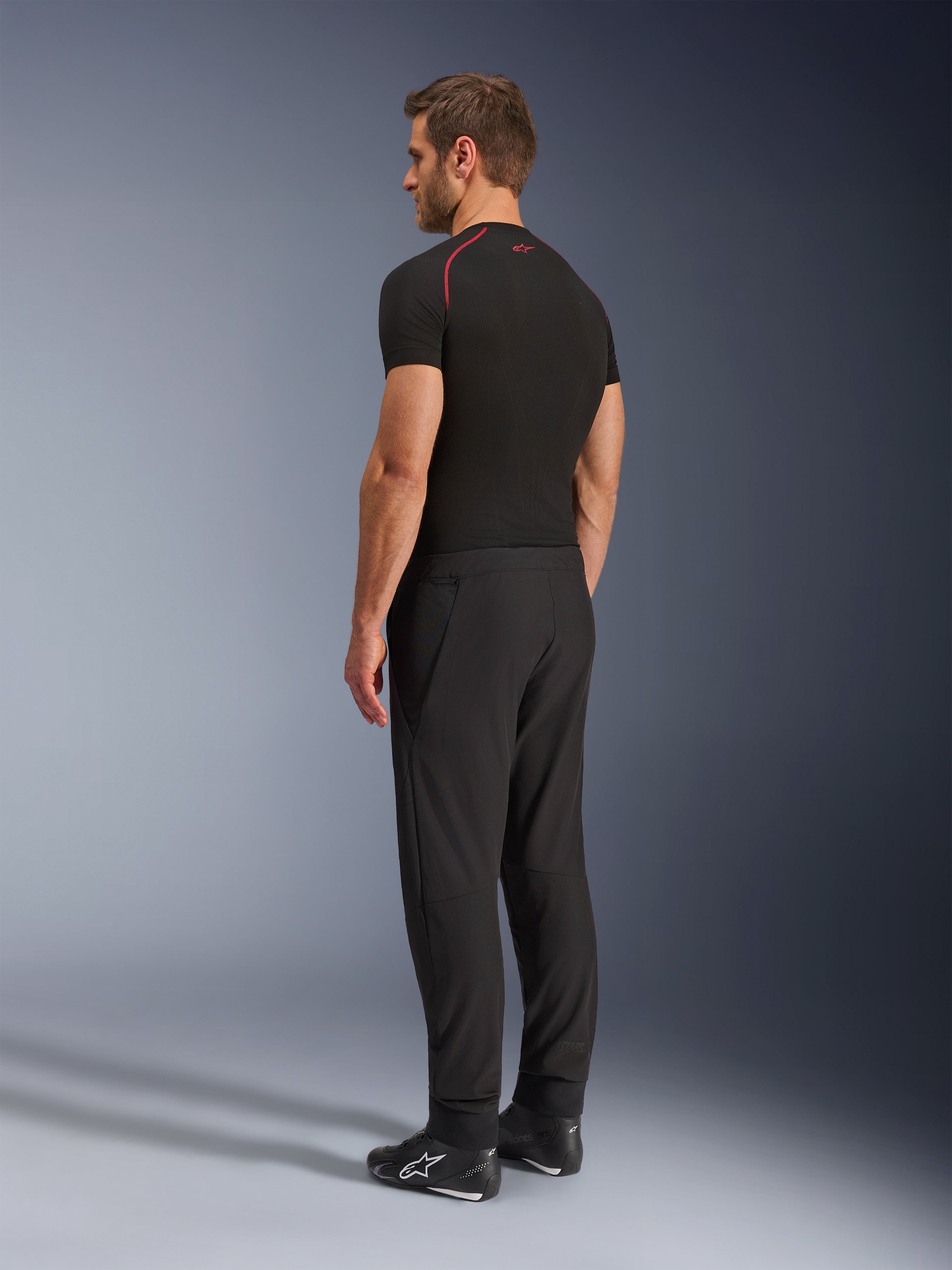 Alpinestars Aeroshell Airflow broek, zwarte motorbroek, lichtgewicht softshell textiel, gedragen met een zwart technisch shirt met korte mouwen en zwarte motorlaarzen, achteraanzicht in profiel toont de slanke pasvorm en ergonomische constructie.