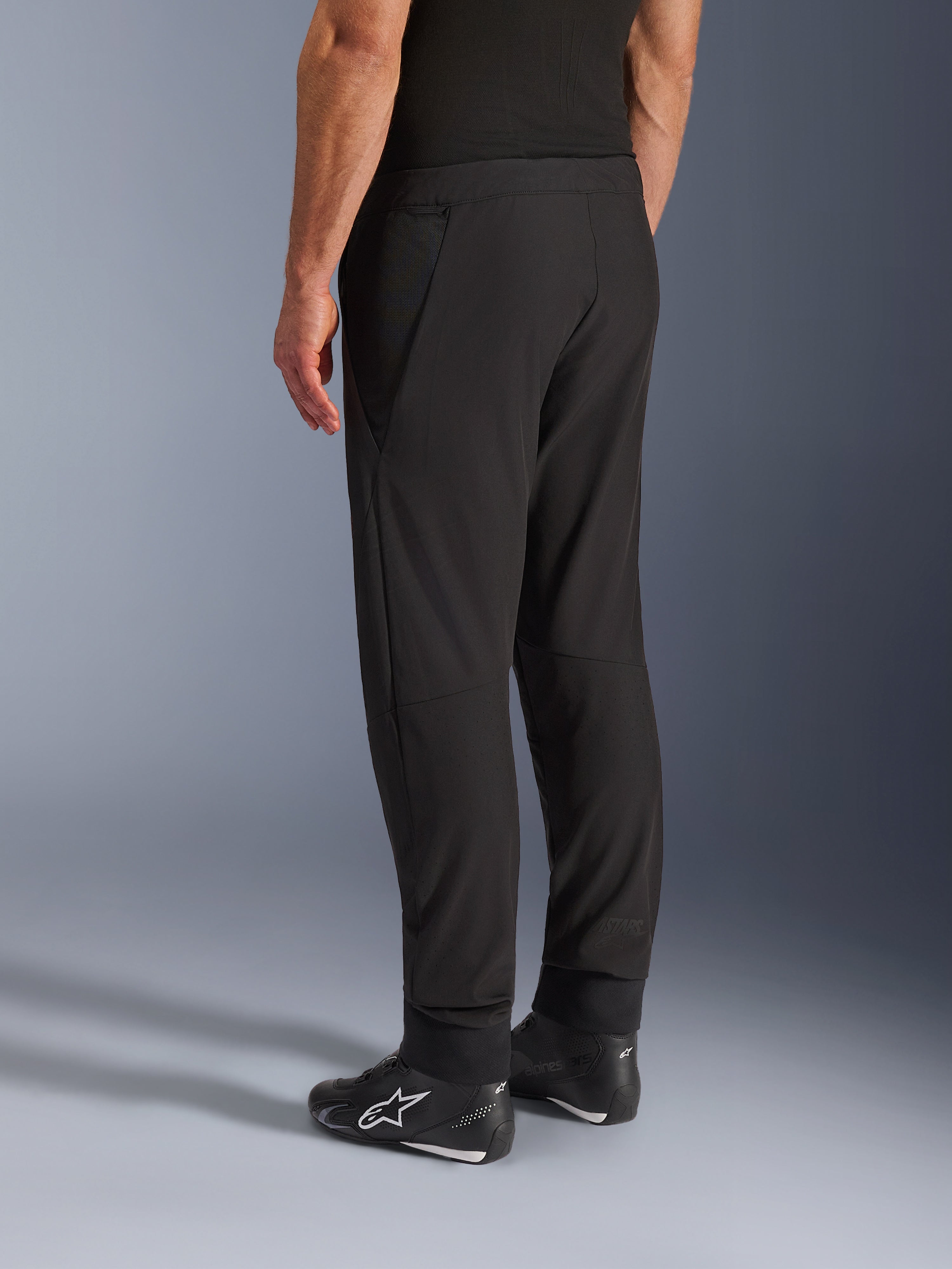 Alpinestars Aeroshell Airflow Pants, zwarte motorbroek, driekwart achteraanzicht, lichtgewicht stretch softshell constructie met geïntegreerde mesh-panelen, verstelbare enkelmanchetten, gedragen met zwarte Alpinestars motorschoenen