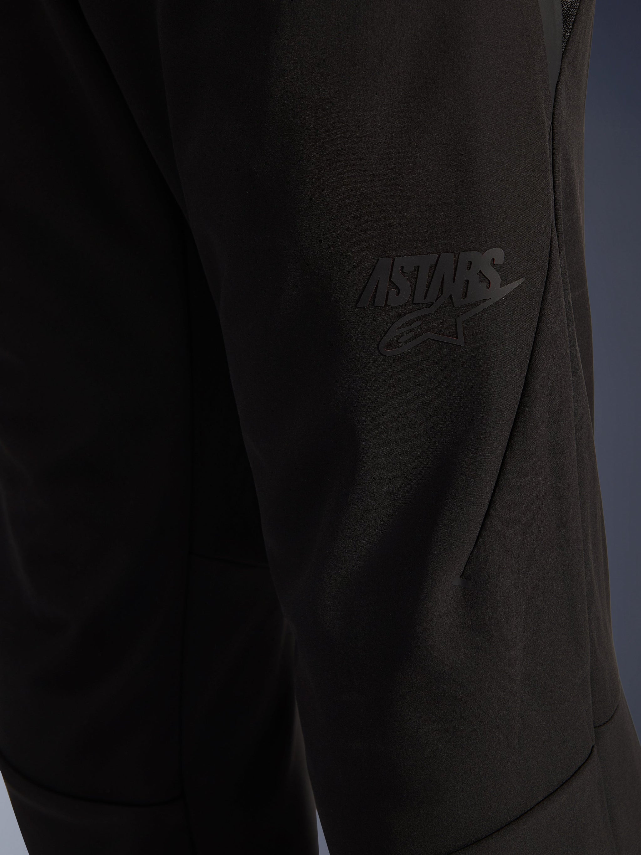 Alpinestars Aeroshell Airflow broek, zwarte motorbroek, close-up van het dijgedeelte met een verhoogd zwart ASTARS-logo, lichtgewicht stretch softshell materiaal met ergonomisch naadontwerp