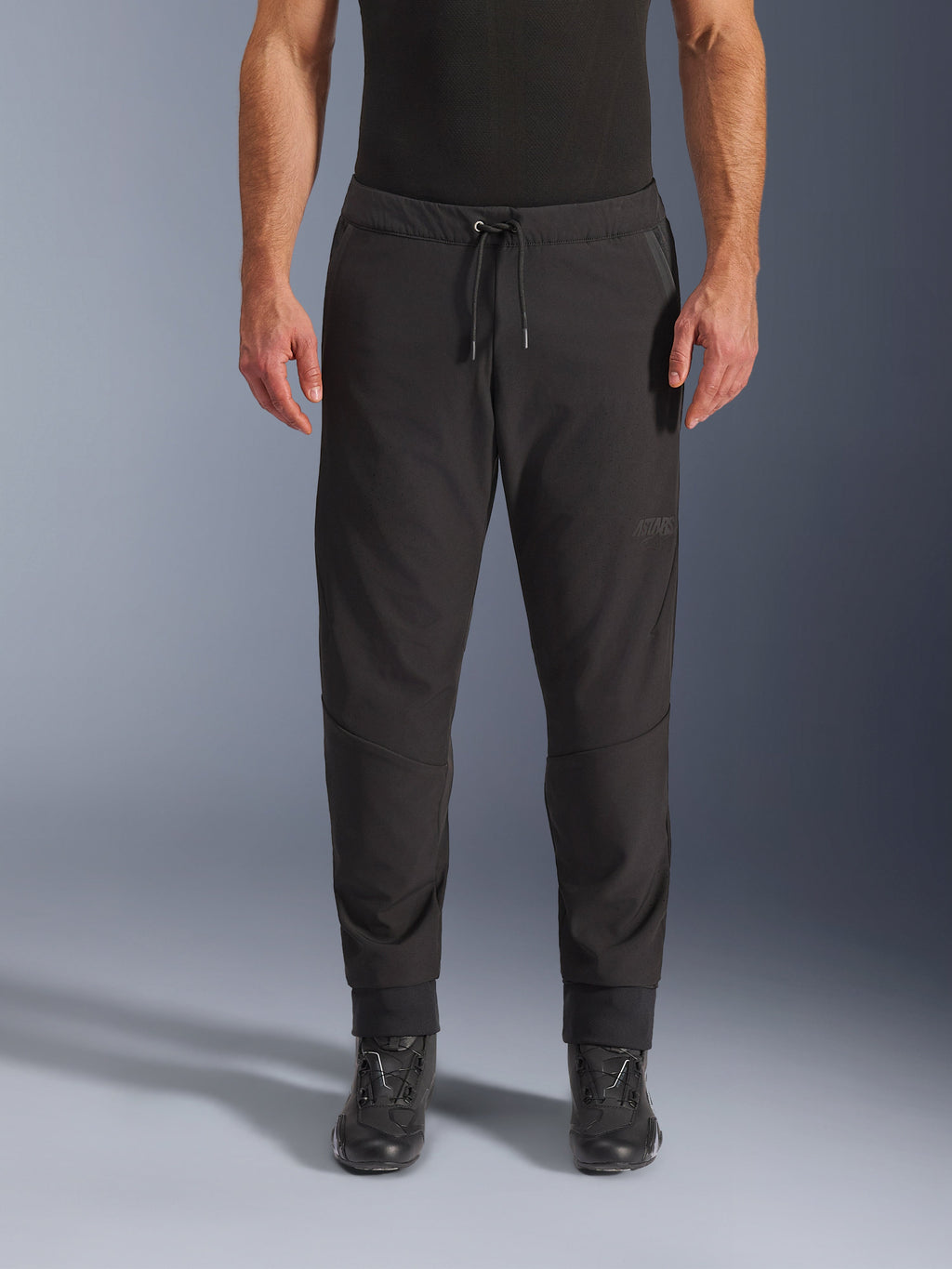 Alpinestars Aeroshell Airflow broek, zwarte motorbroek, vooraanzicht gedragen met zwarte motorlaarzen, voorzien van een taille met trekkoord en taps toelopende enkelmanchetten, lichtgewicht softshell-ontwerp met subtiele ton-sur-ton branding.