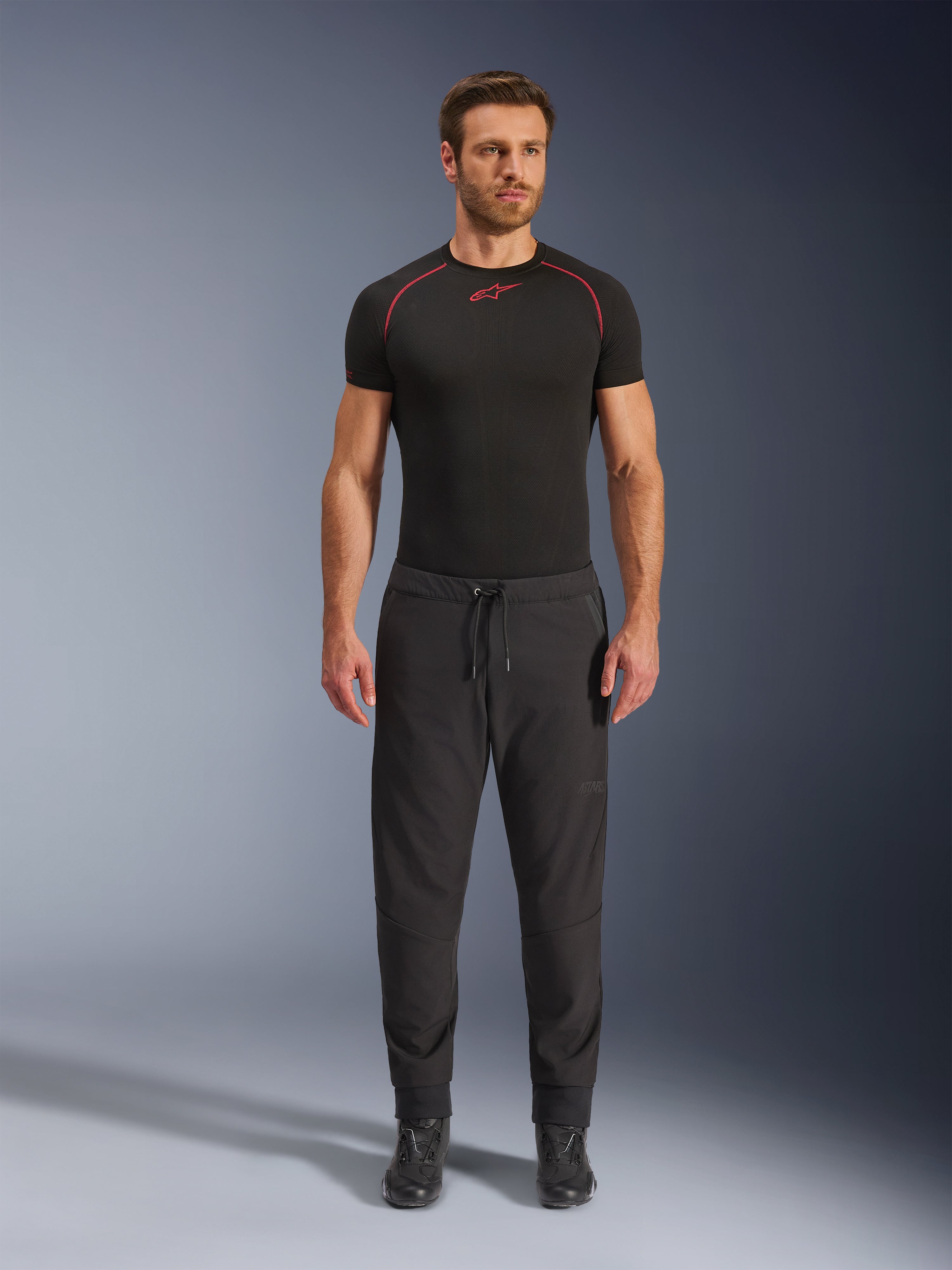 Model draagt Alpinestars Aeroshell Airflow Broek in het zwart, gecombineerd met een zwarte basislaag met korte mouwen en zwarte motorlaarzen. Deze lichtgewicht softshell motorbroek is voorzien van een taille met trekkoord en elastische manchetten, ontworpen voor sport- en toerrijders in warme weersomstandigheden.