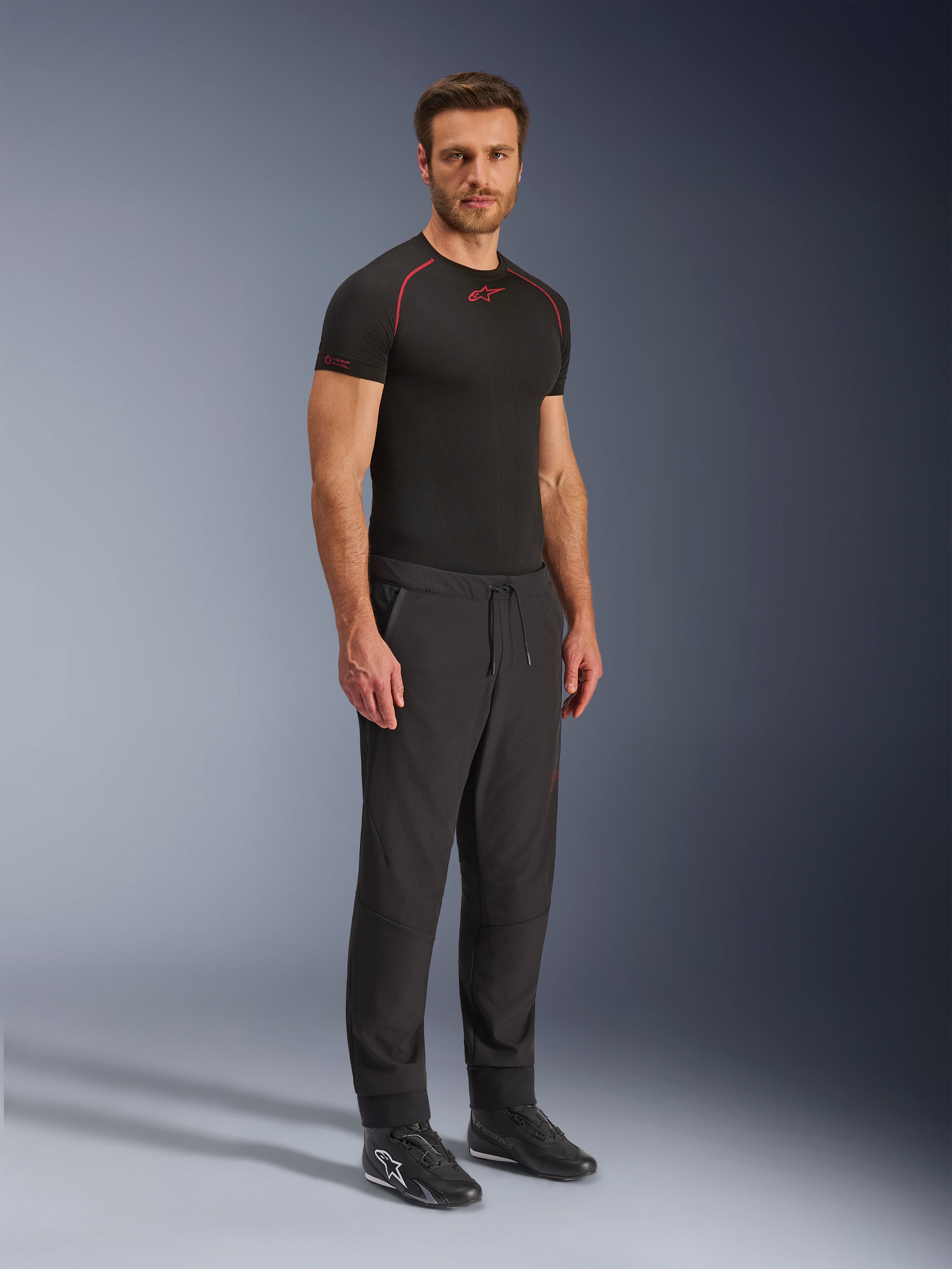 Alpinestars Aeroshell Airflow Broek, zwarte motorbroek, volledige studiofoto van een model die een softshell broek draagt met een trekkoord in de taille en elastische boorden, gecombineerd met een zwart Alpinestars base layer shirt en zwarte motorschoenen.