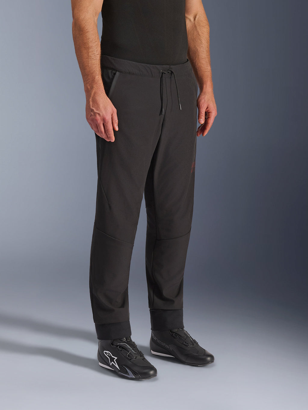 Alpinestars Aeroshell Airflow broek, zwarte motorbroek, gedragen door model met zwarte rijschoenen, voorzien van een tailleband met trekkoord, nauwsluitende enkelboorden en laser-geperforeerde softshell-stof voor maximale ventilatie