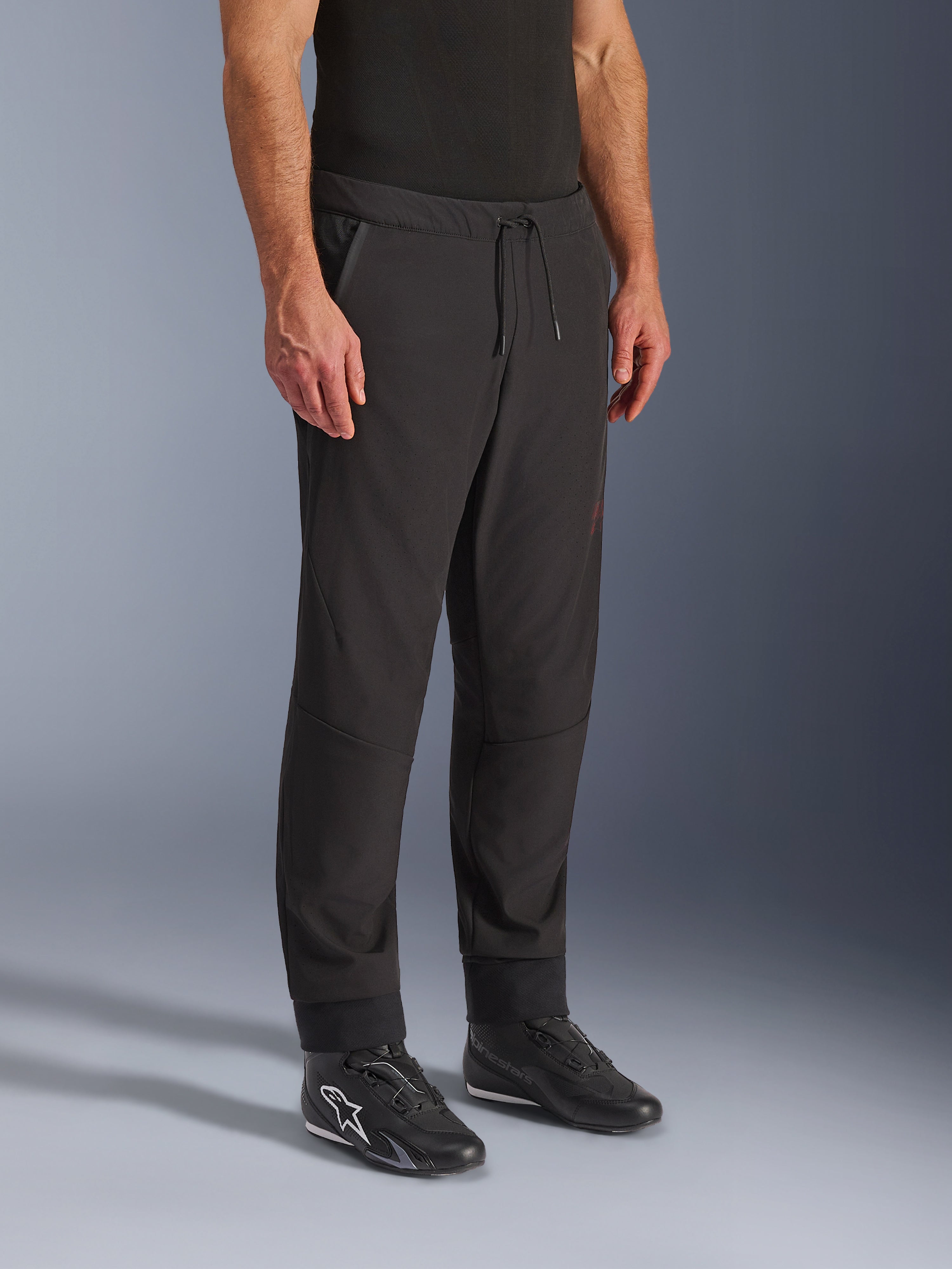 Alpinestars Aeroshell Airflow broek, zwarte motorbroek, gedragen door model met zwarte rijschoenen, voorzien van een tailleband met trekkoord, nauwsluitende enkelboorden en laser-geperforeerde softshell-stof voor maximale ventilatie