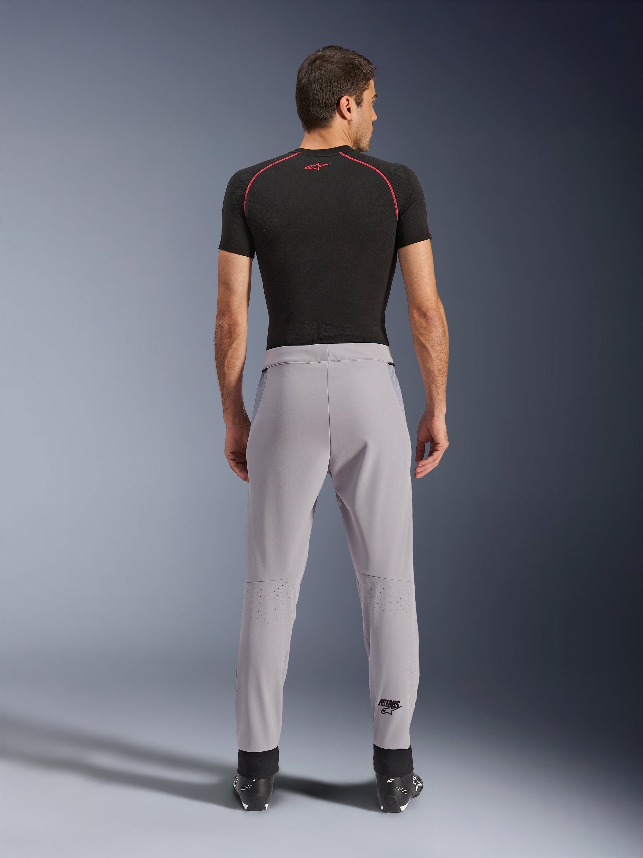 Alpinestars Aeroshell Airflow Broek, Mid Gray, achteraanzicht met laser-geperforeerde zones op de kuiten, lichtgewicht stretch softshell constructie, gedragen met een zwart shirt met korte mouwen en motorlaarzen, sportieve pasvorm voor rijden in warm weer